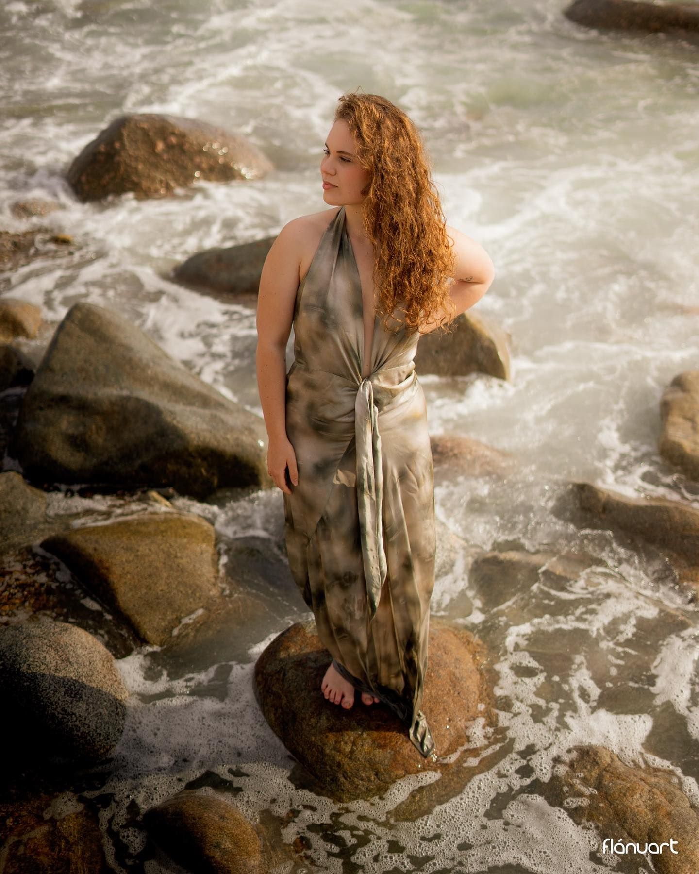 Redhead woman in tie-dye halter dress standing on coastal rocks surrounded by crashing ocean waves.