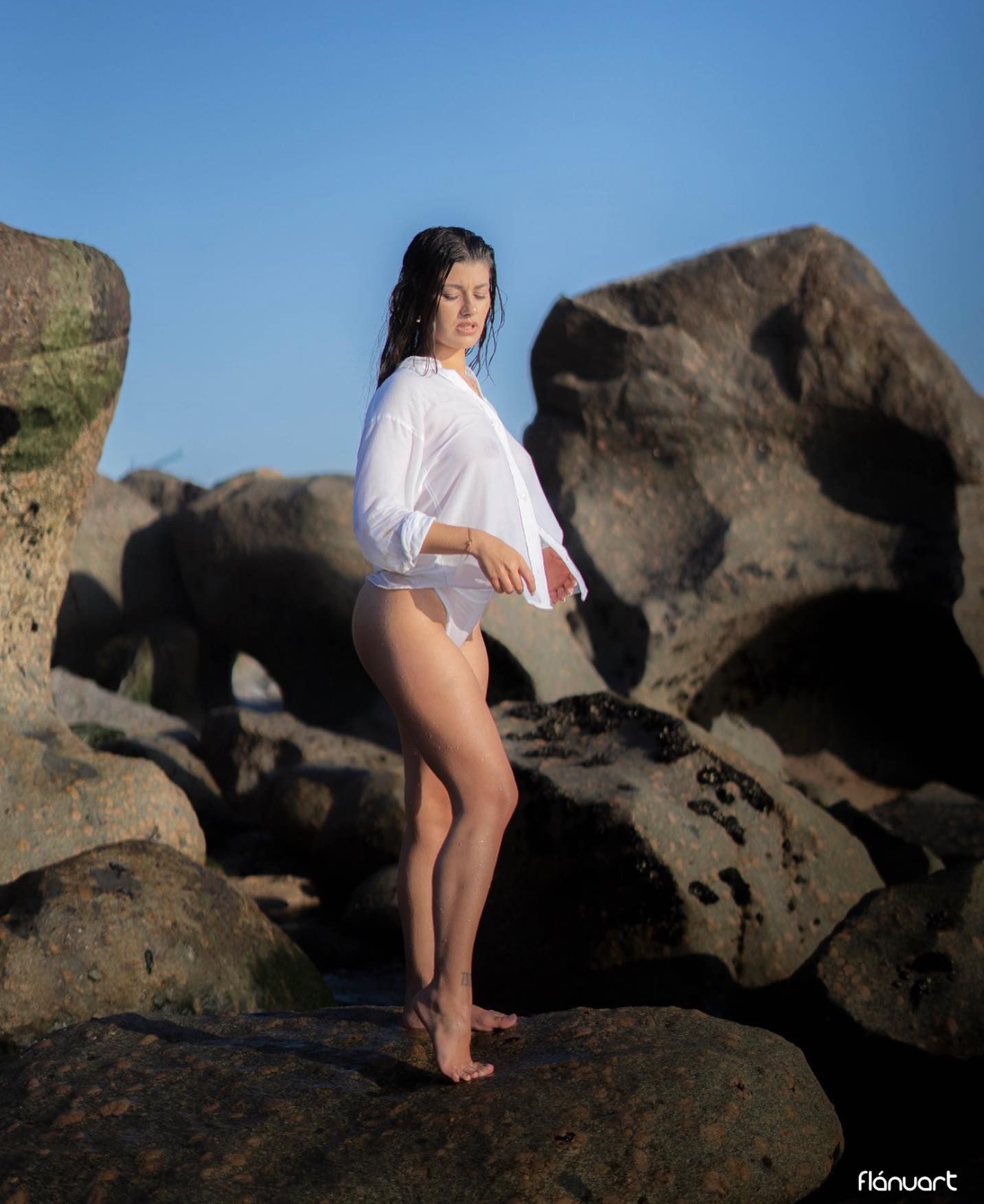 Brunette woman in white shirt and underwear posing on large coastal rocks under blue sky.