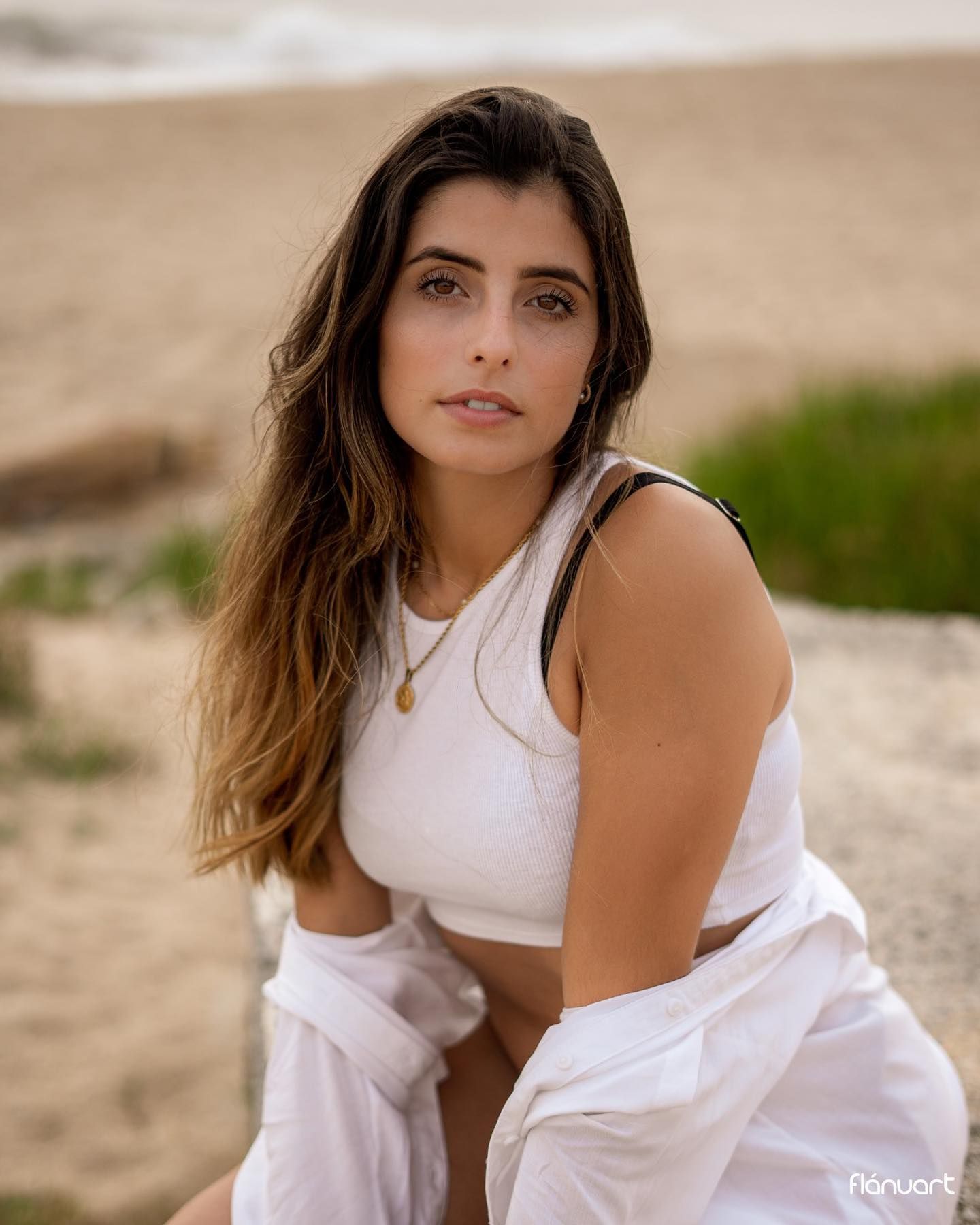 Young woman in white crop top and pants sits on a sandy beach with natural wavy hair.