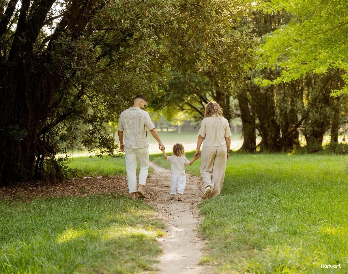 Family of three walking hand-in-hand down a sunny park path surrounded by lush green trees.