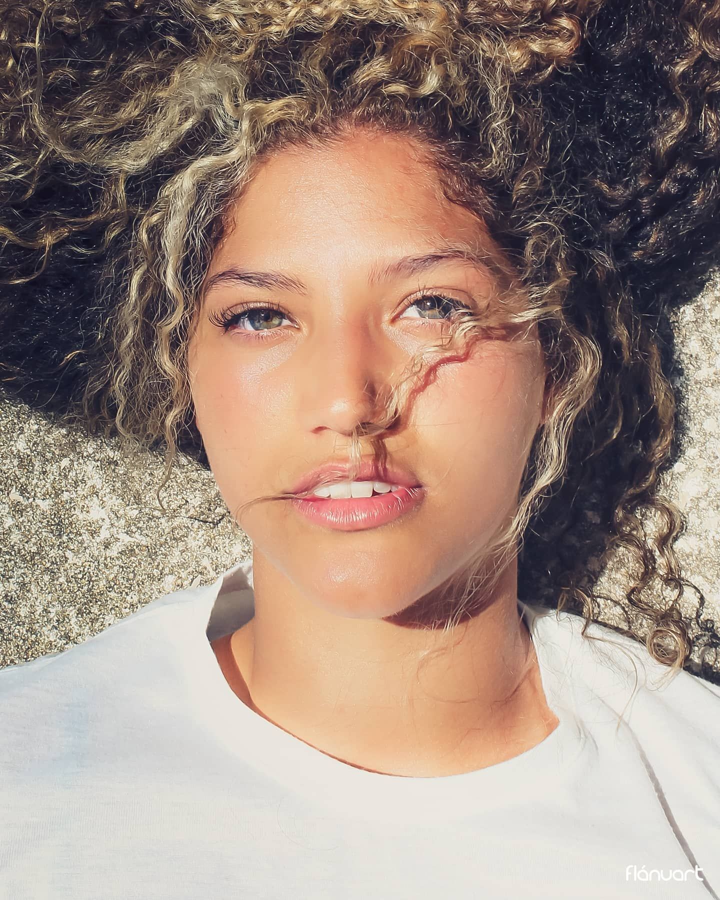 Close-up portrait of a young woman with curly highlighted hair lying in sunlight outdoors.