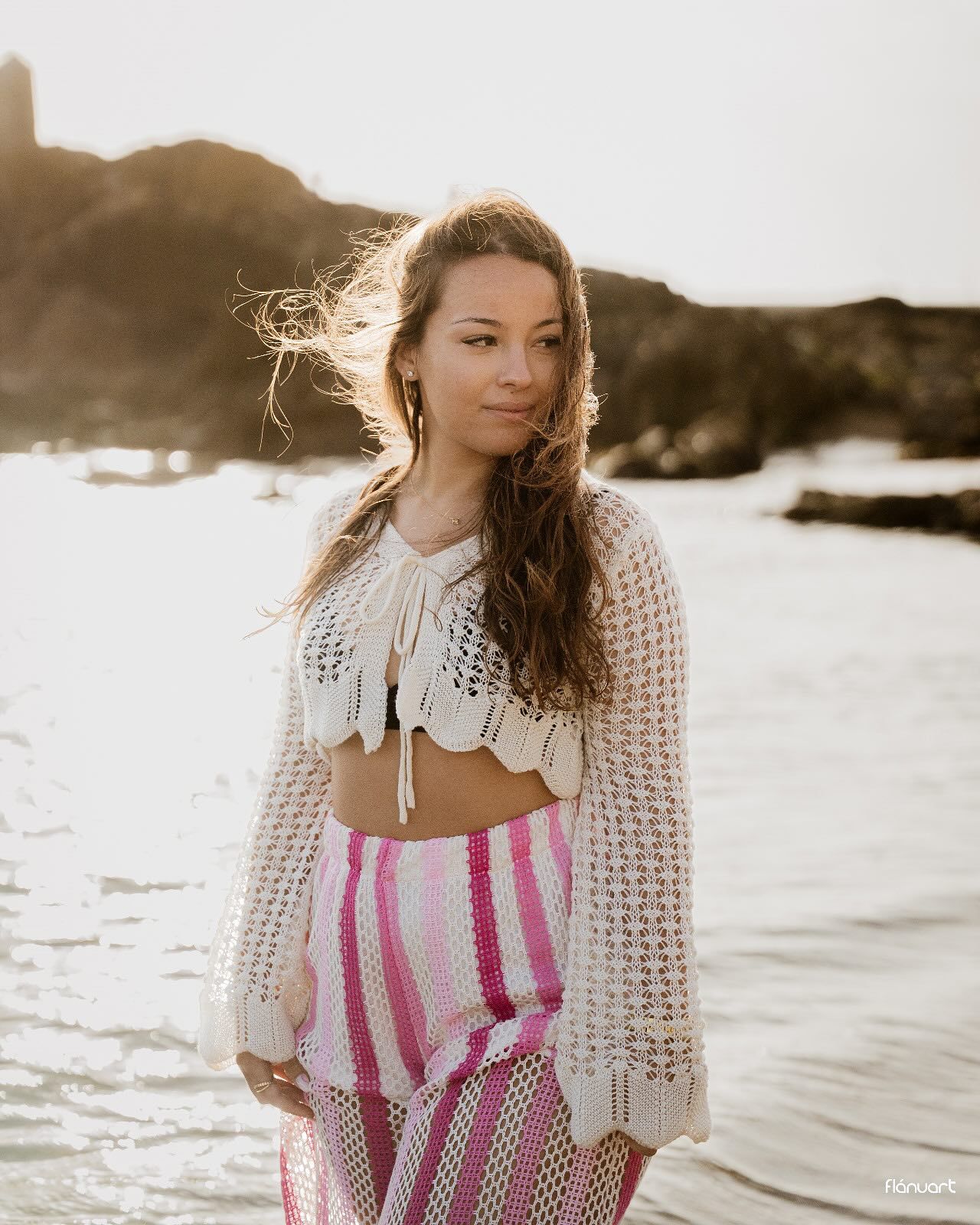 Young woman in crochet top and pink striped skirt standing on a beach at golden hour sunset.