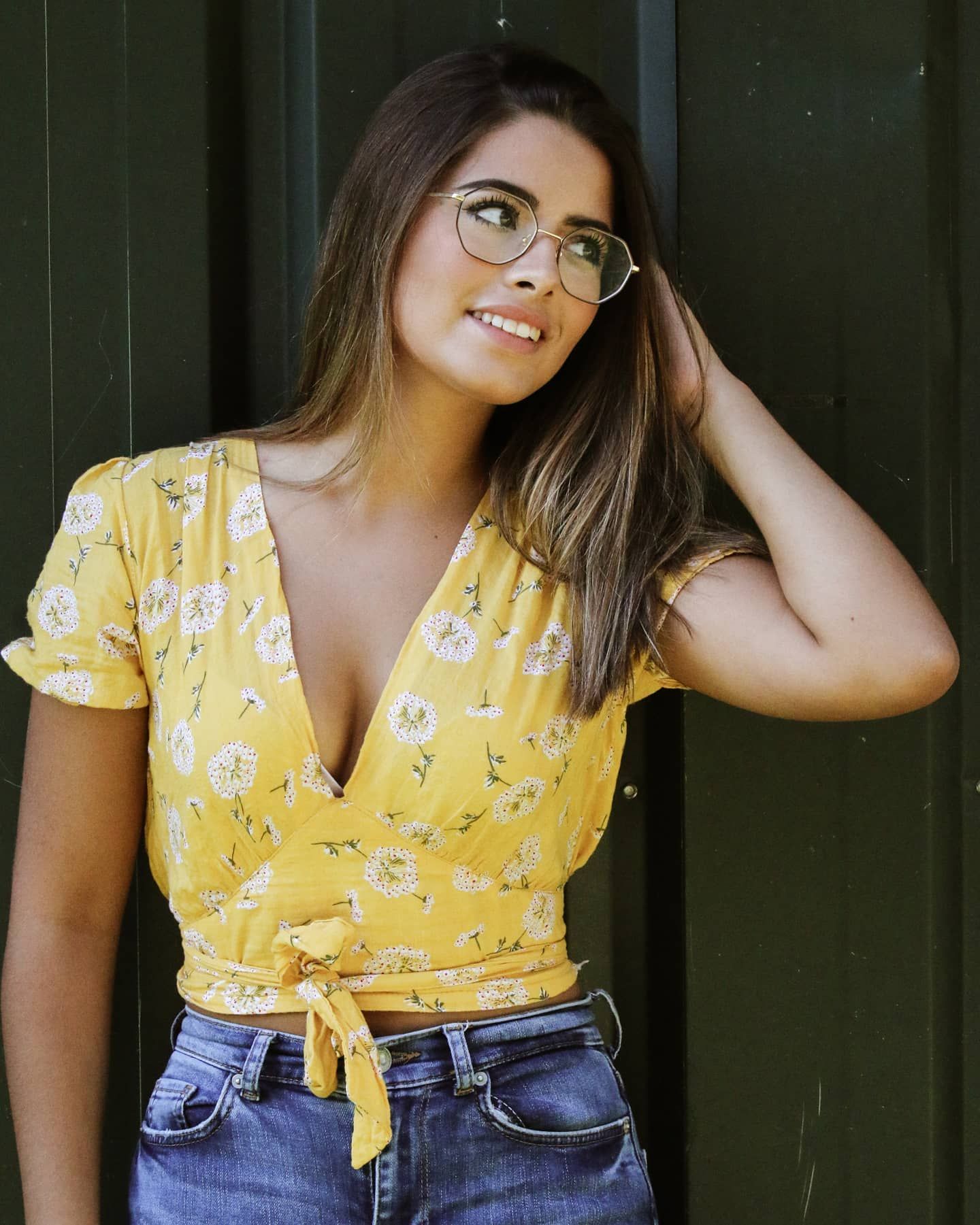 Smiling woman in yellow floral wrap crop top and jeans wearing round glasses outdoors.