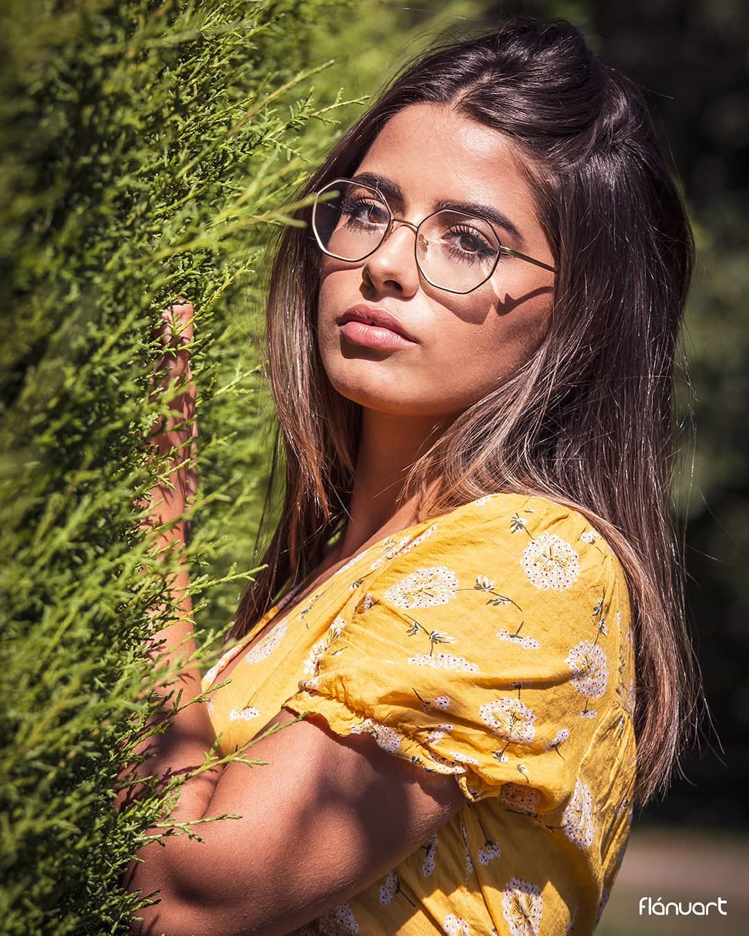 Young woman with glasses in yellow floral dress posing outdoors near green hedge in sunlight.