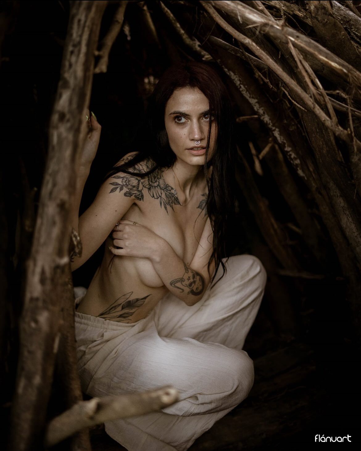 Tattooed redhead woman posing topless inside a rustic wooden shelter with dramatic moody lighting.