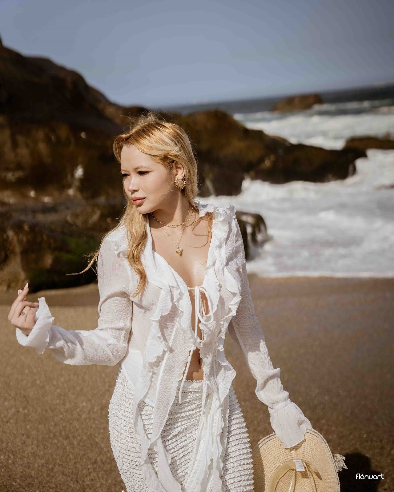Blonde woman in white ruffled crop top and skirt walking on rocky beach holding a straw bag.