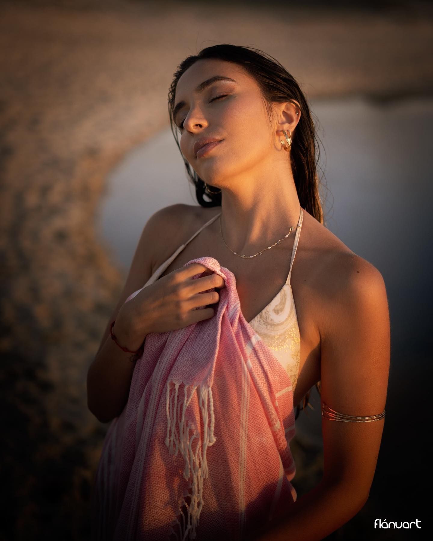 Woman with wet hair wrapped in pink shawl posing on beach in warm golden sunset light.
