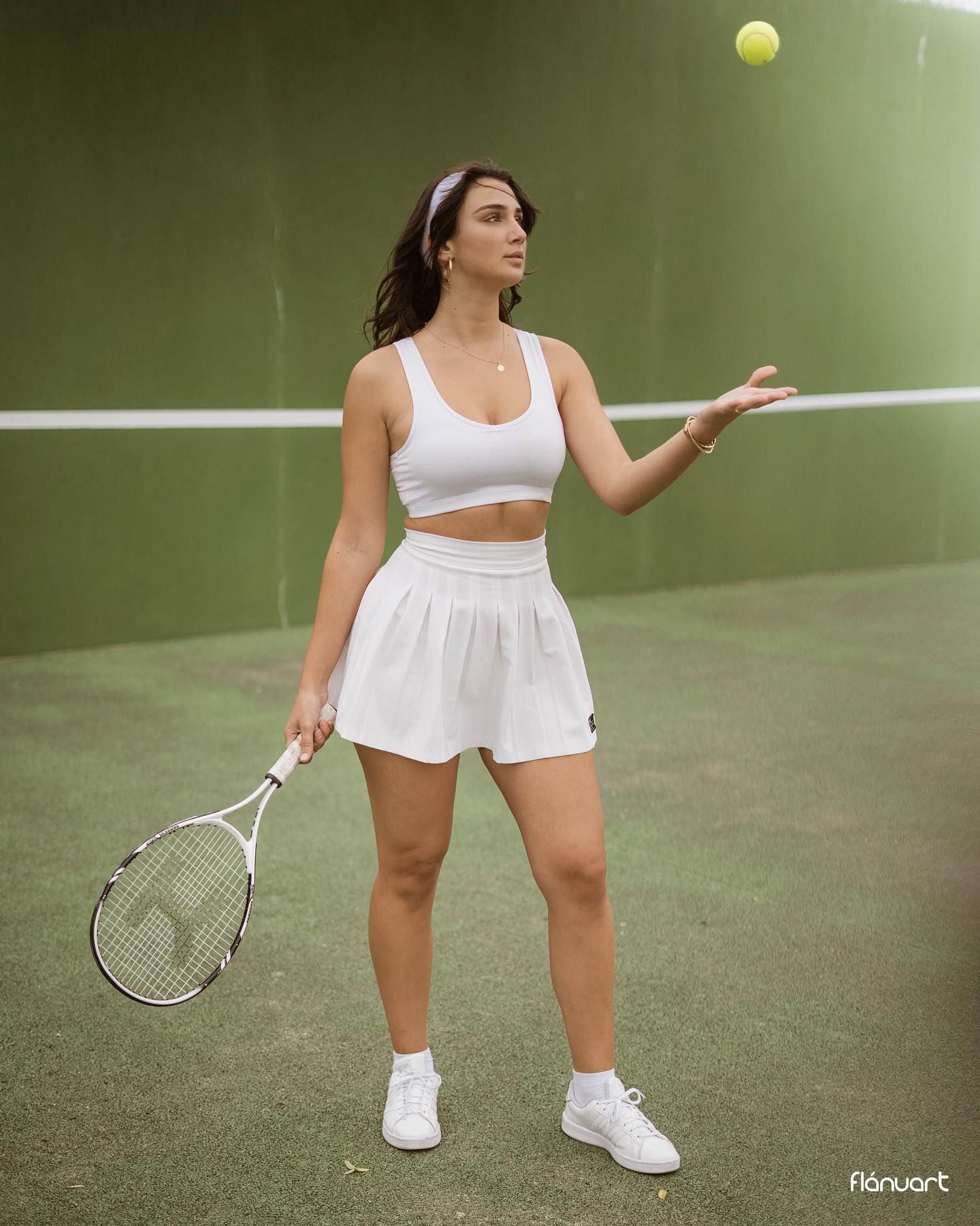 Woman in white tennis outfit tosses ball while holding racket on green court.