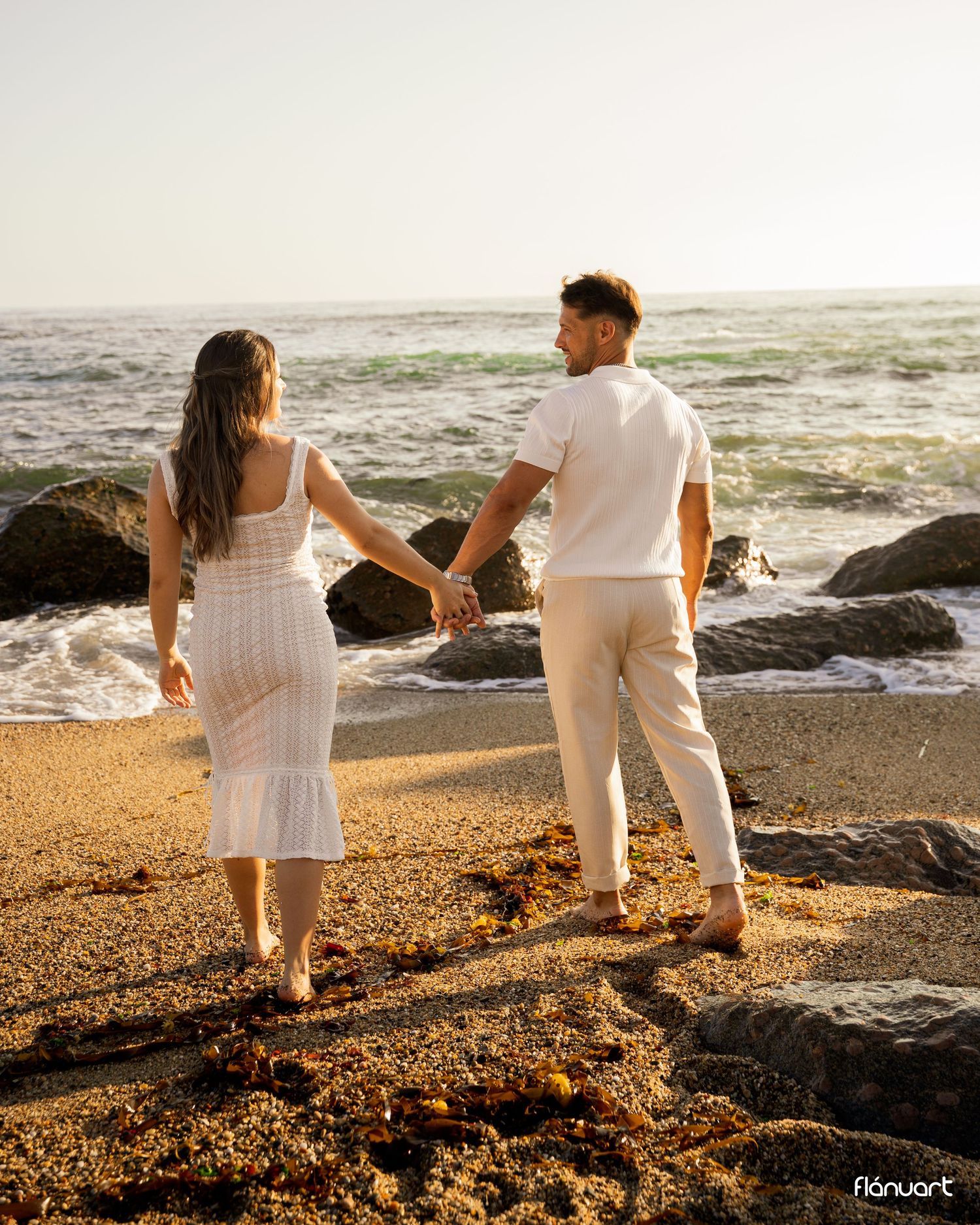 Couple holding hands walking on a rocky beach at golden hour, dressed in matching white outfits.