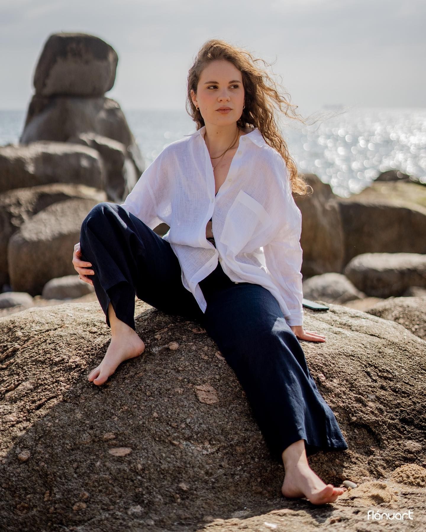 Young woman in white shirt and navy pants sitting barefoot on coastal rocks with ocean in background.