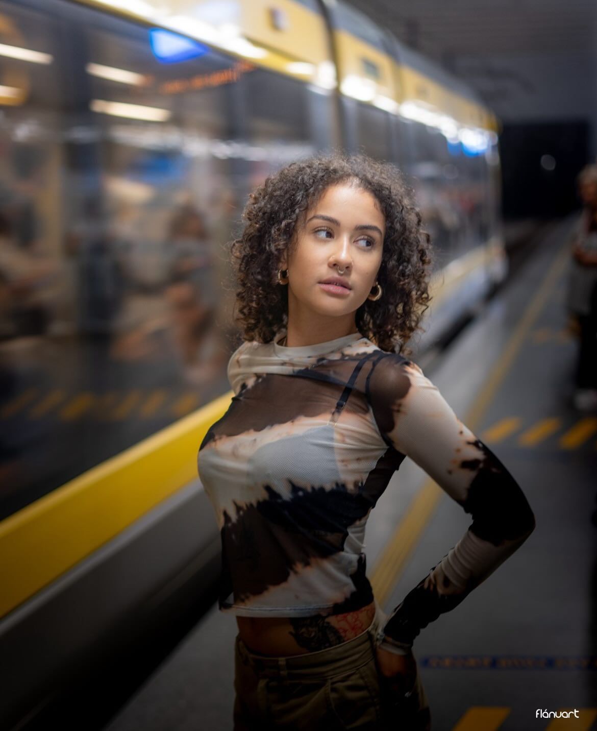Young woman with curly hair in tie-dye mesh top posing at a subway station with yellow train in background.