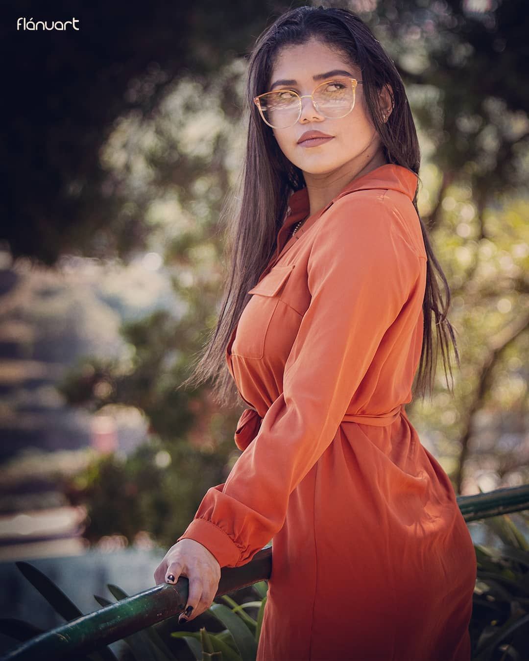 Young woman in an orange belted dress and clear glasses posing outdoors with bokeh background.