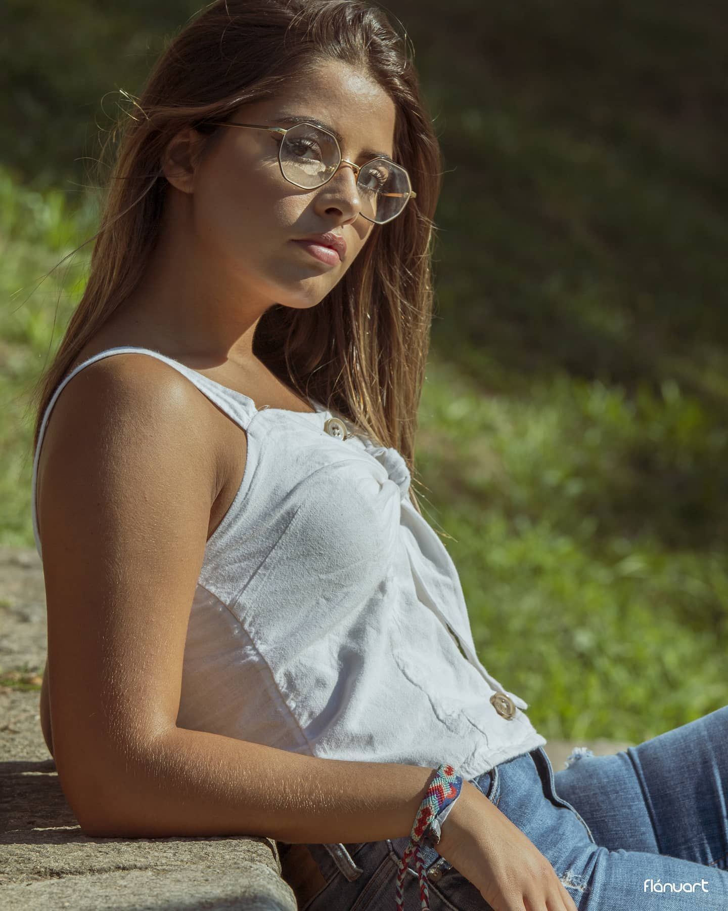 Young woman in white tank top and jeans wearing round glasses sitting outdoors in sunlight.