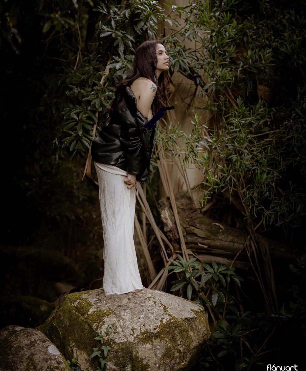 Woman in black jacket and white skirt stands on mossy rock in lush forest.