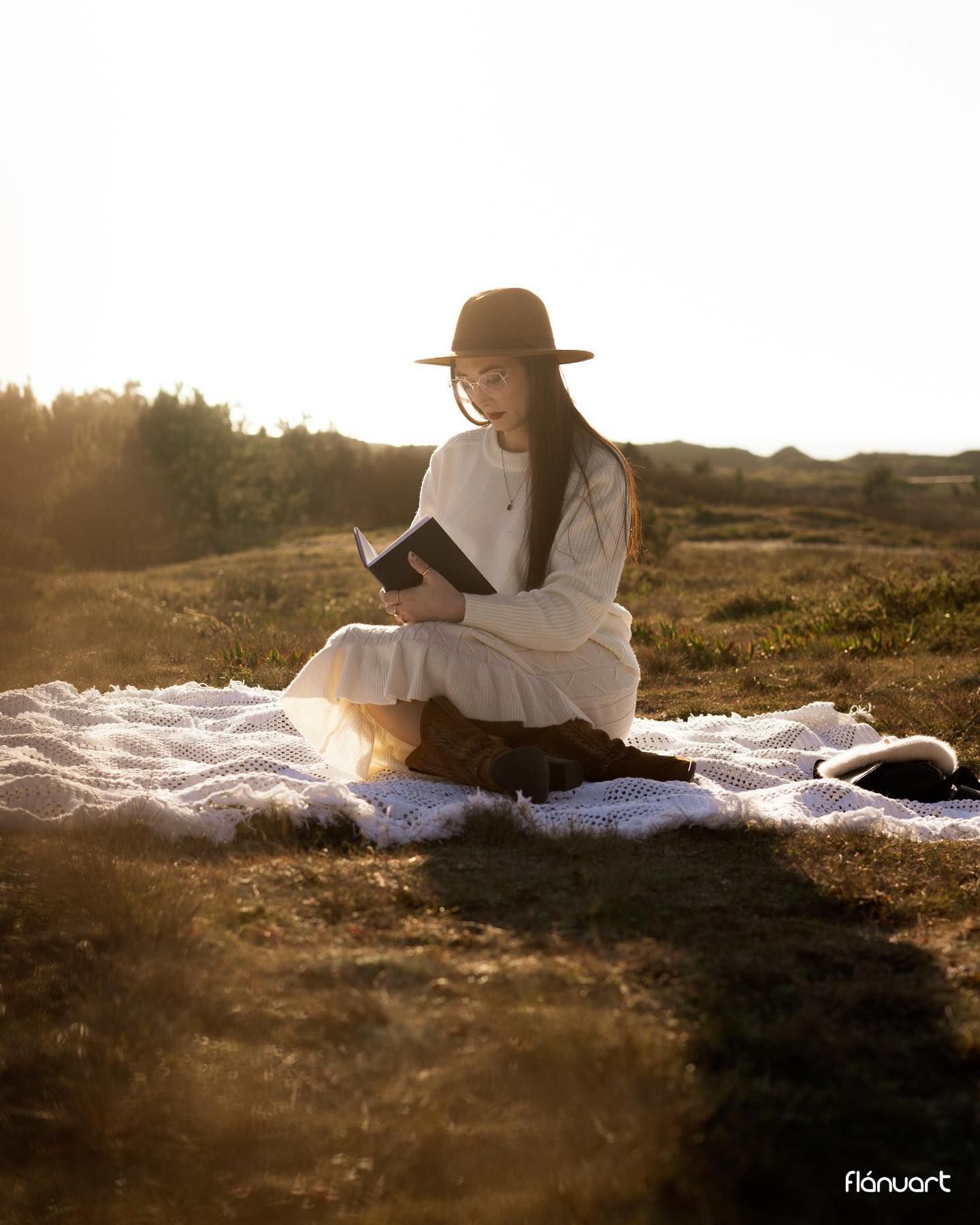 Woman in white outfit and hat sitting on blanket reading book in sunlit golden field.