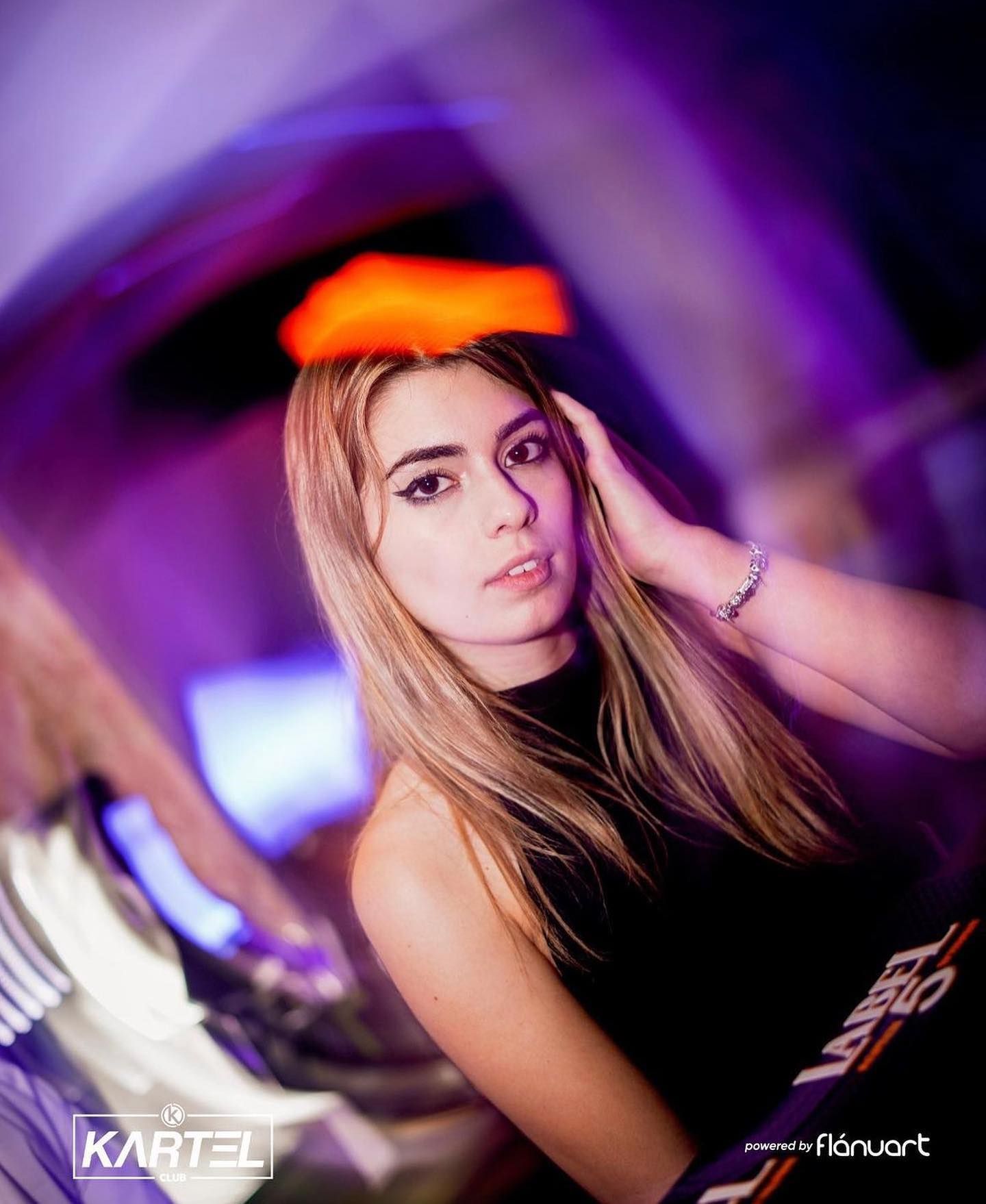Young blonde woman posing at Kartel Club nightclub with purple ambient lighting in the background.