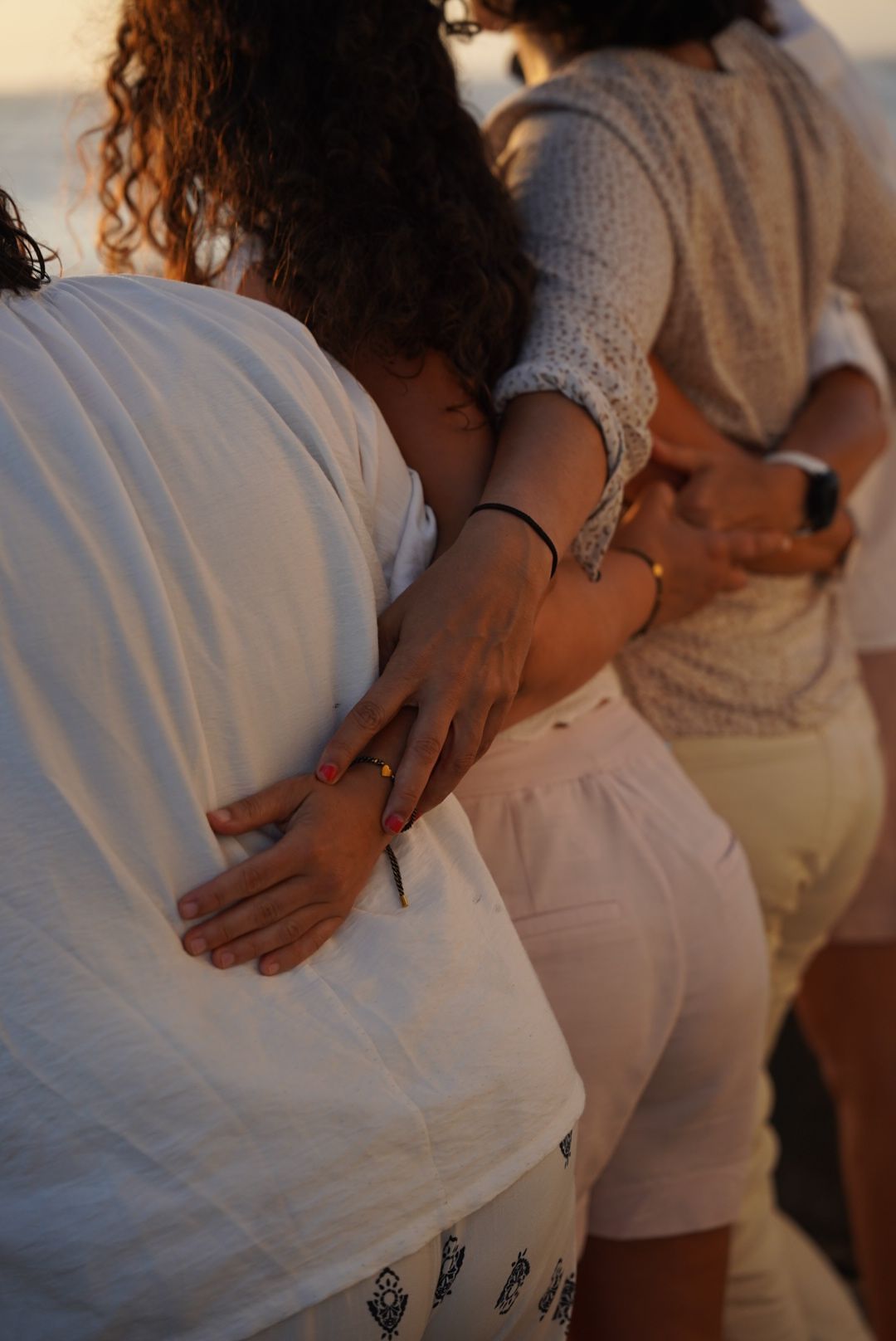 Close-up of people embracing each other from behind on a beach during sunset, wearing white outfits.