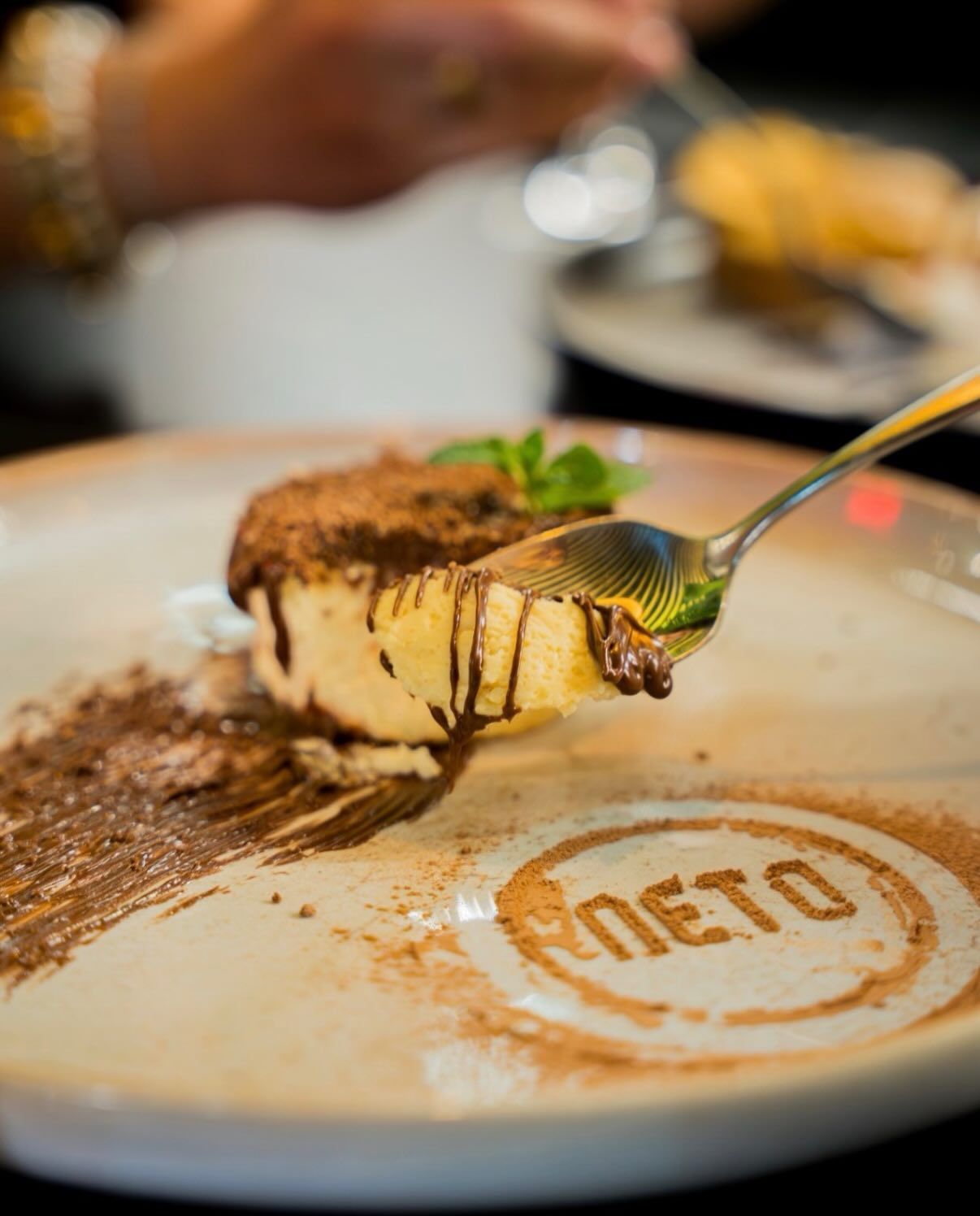 Close-up of a tiramisu dessert on a branded Neto plate with a spoon lifting a bite.