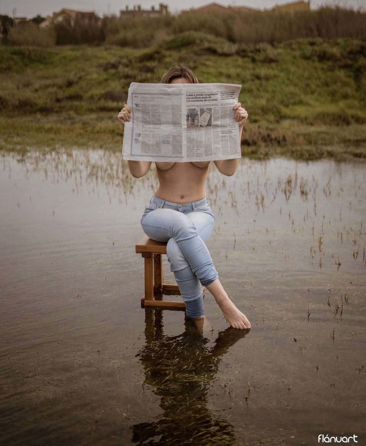 Topless woman sitting on a stool in water reading a newspaper in an outdoor art photo.