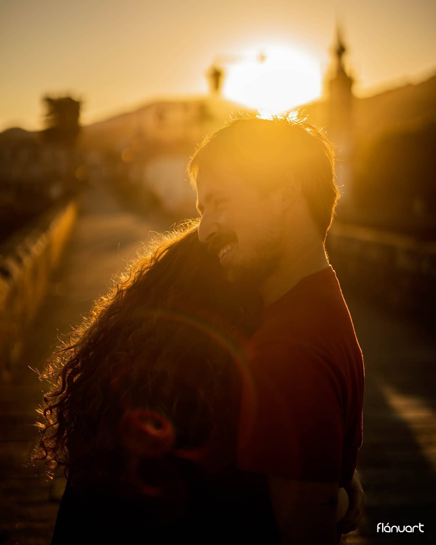 Romantic couple embracing at sunset on a bridge with warm golden backlight and city skyline.