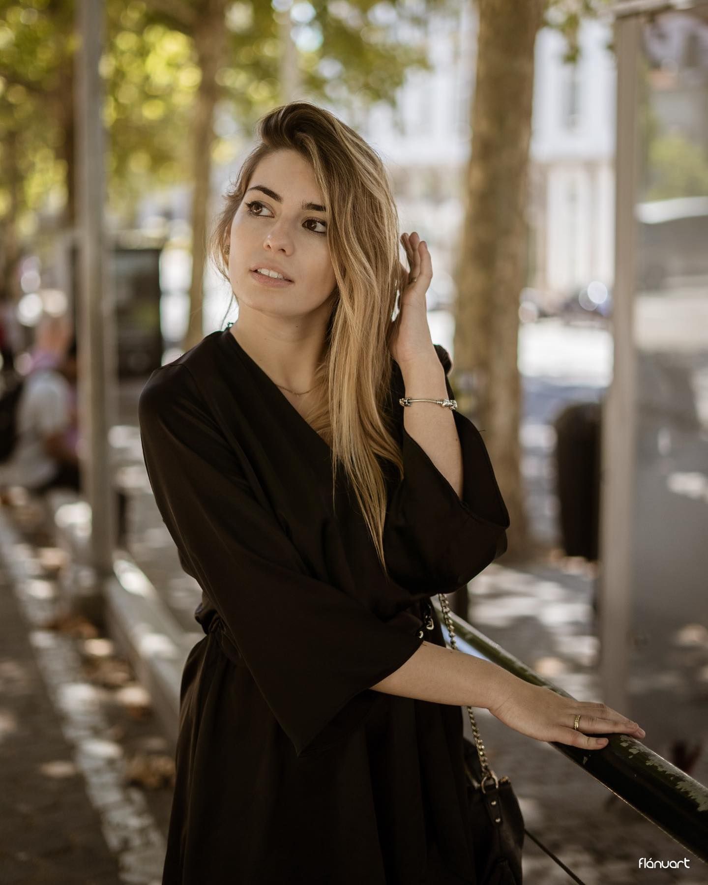 Blonde woman in black wrap dress leaning on railing on a tree-lined urban sidewalk.