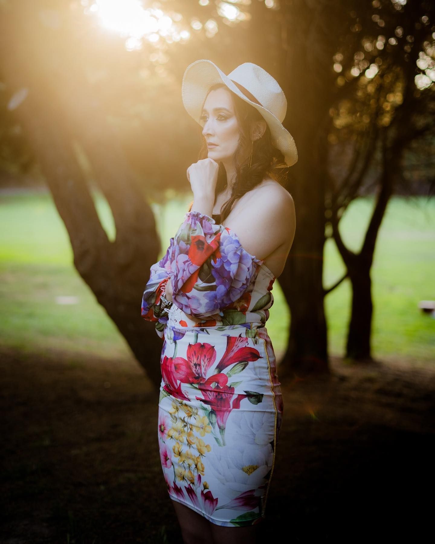 Woman in floral off-shoulder dress and white hat posing in golden sunset light among trees.