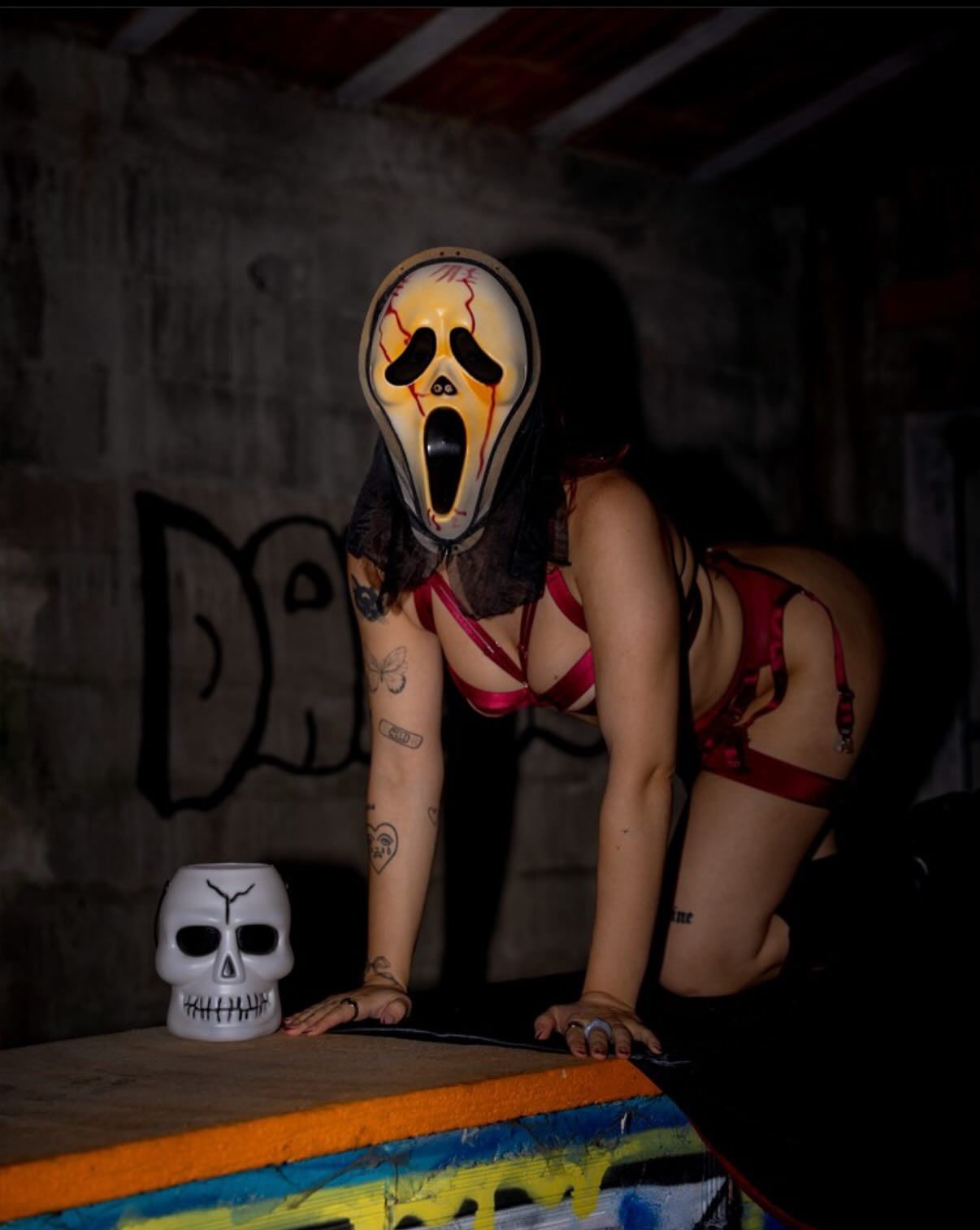 Woman wearing painted Ghostface mask in red lingerie posing in dark graffiti room with skull prop.
