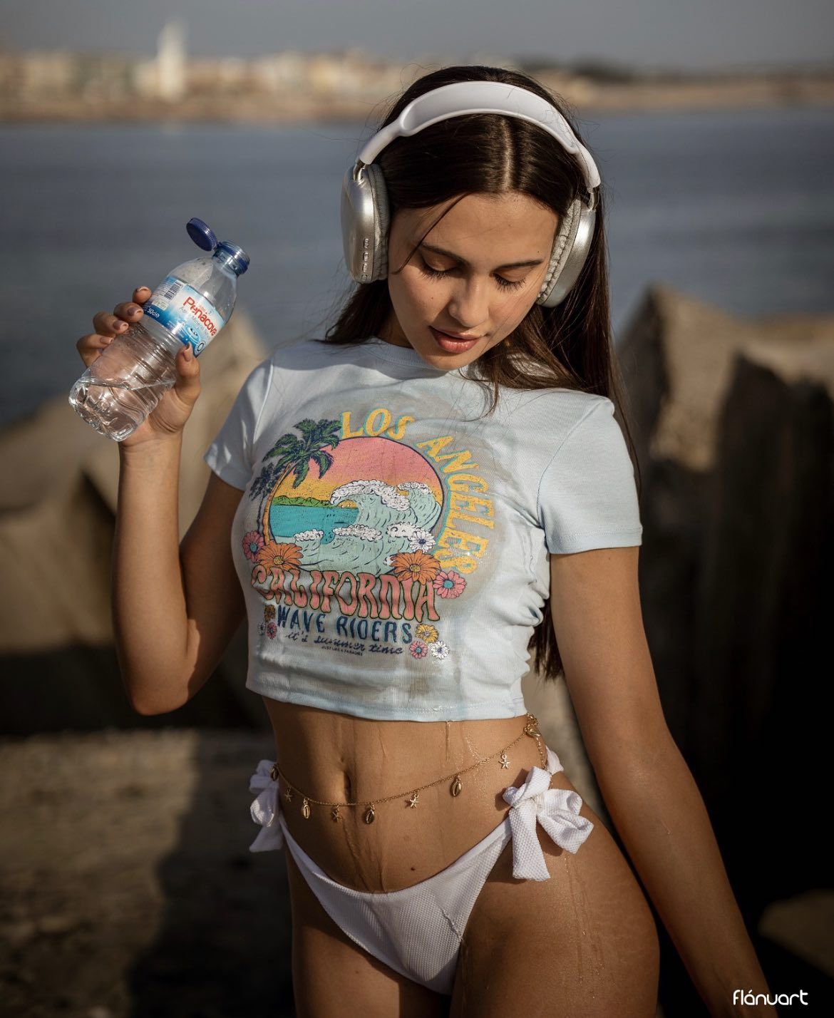 Young woman in crop tee and white bikini bottom holding water bottle near rocky shore.