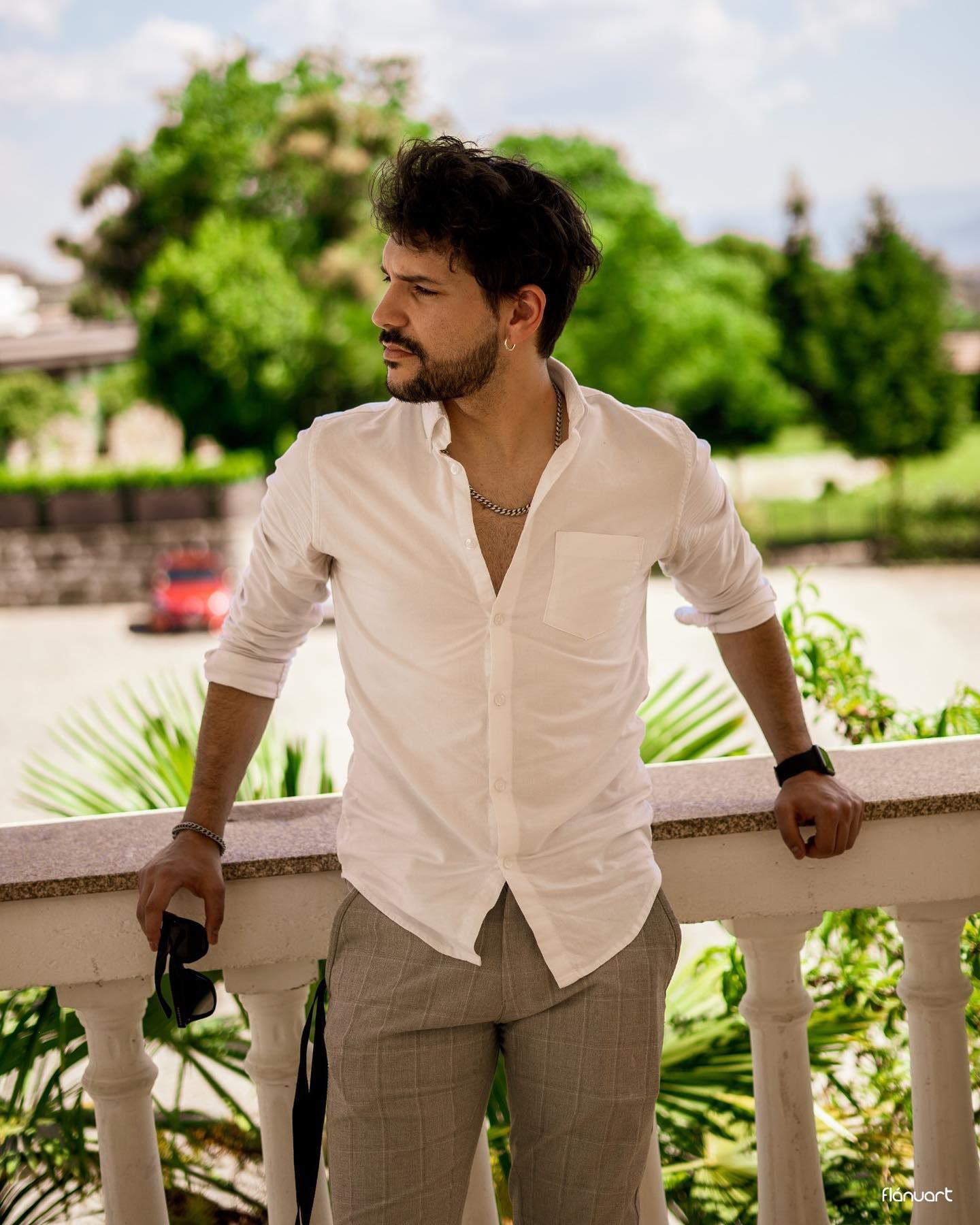 Stylish man in white linen shirt leaning on a white balustrade in a lush garden.
