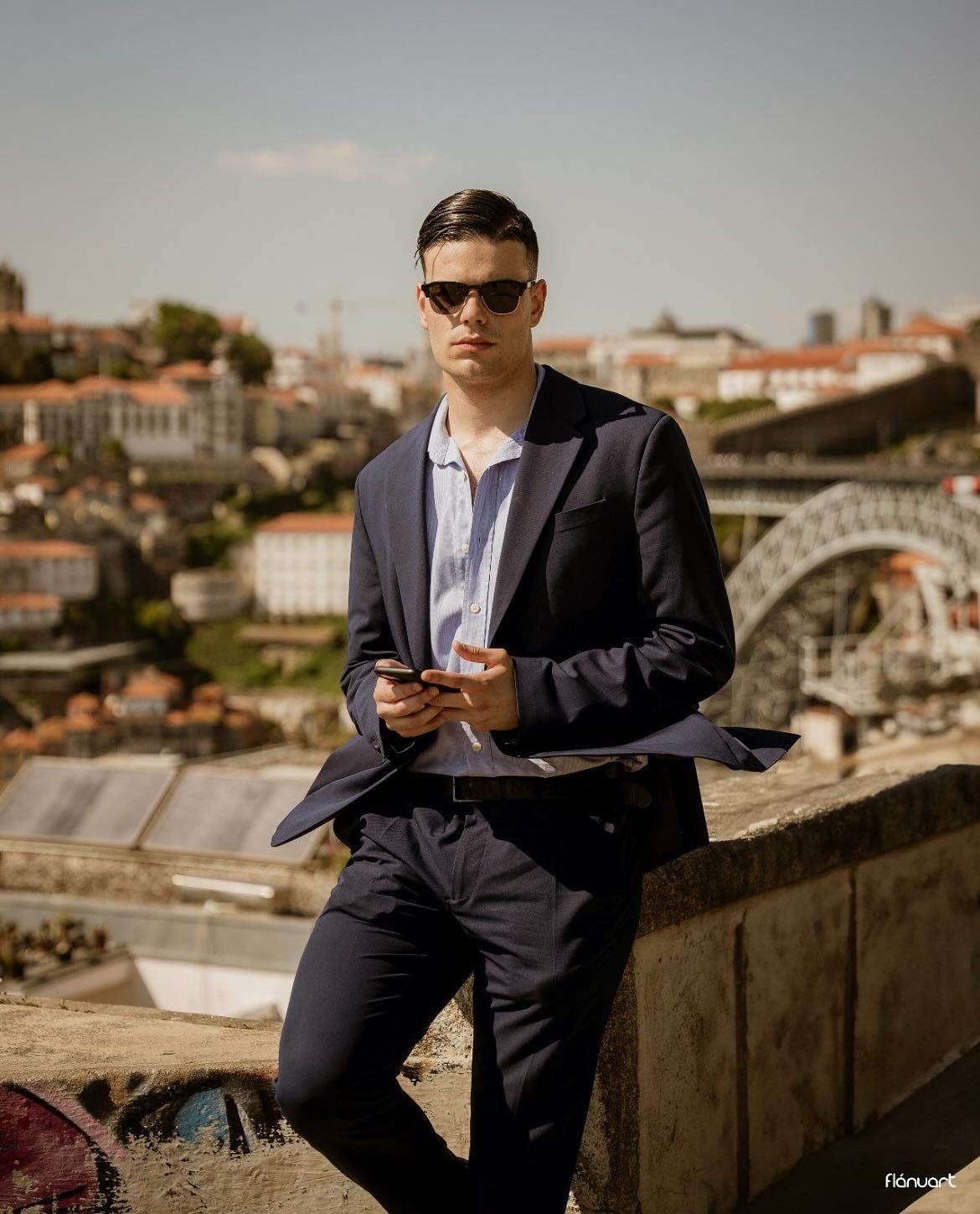 Stylish young man in navy blue suit with sunglasses leaning on a wall with a bridge cityscape behind him.