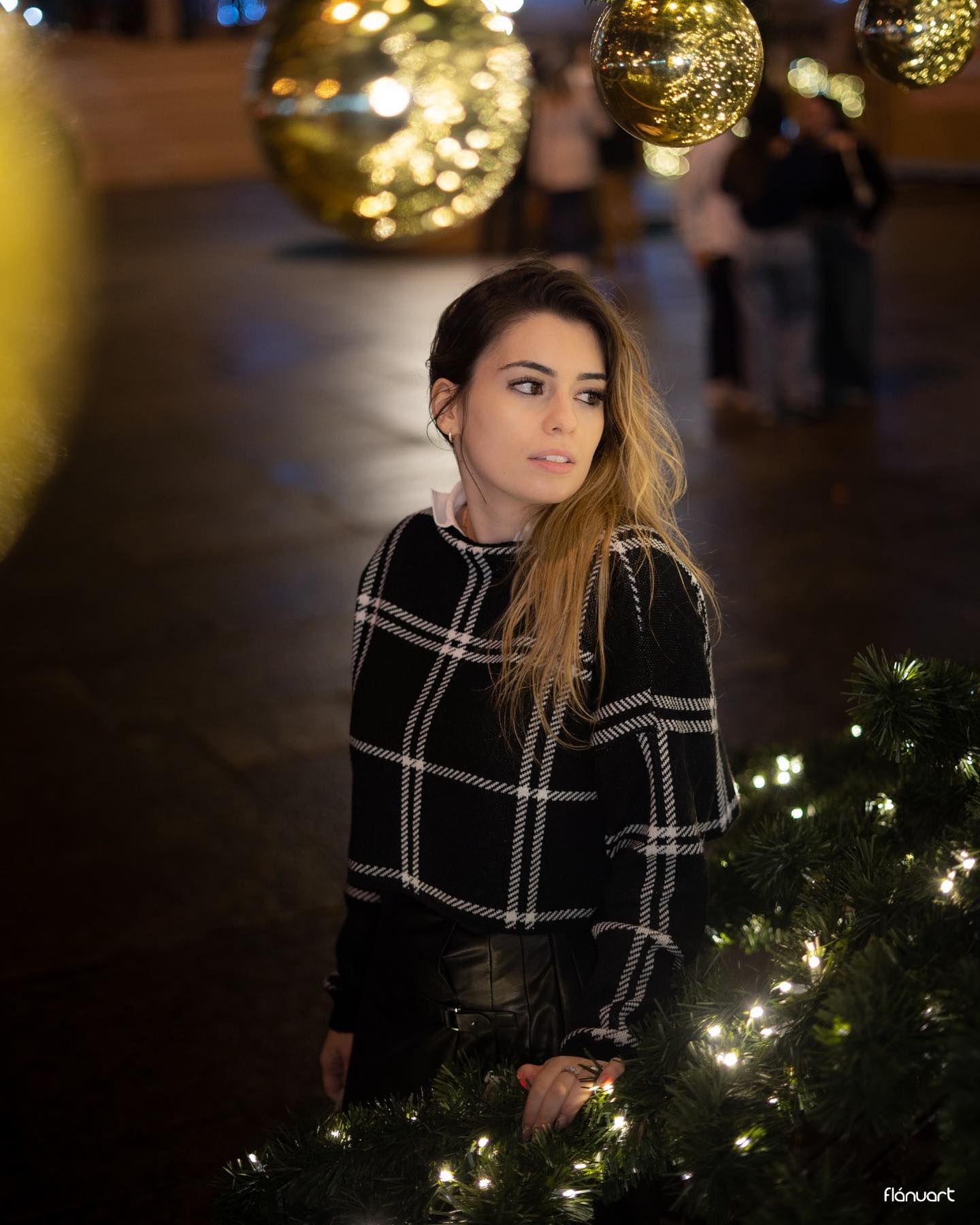 Young woman in plaid sweater posing near Christmas tree with golden bokeh lights at night.