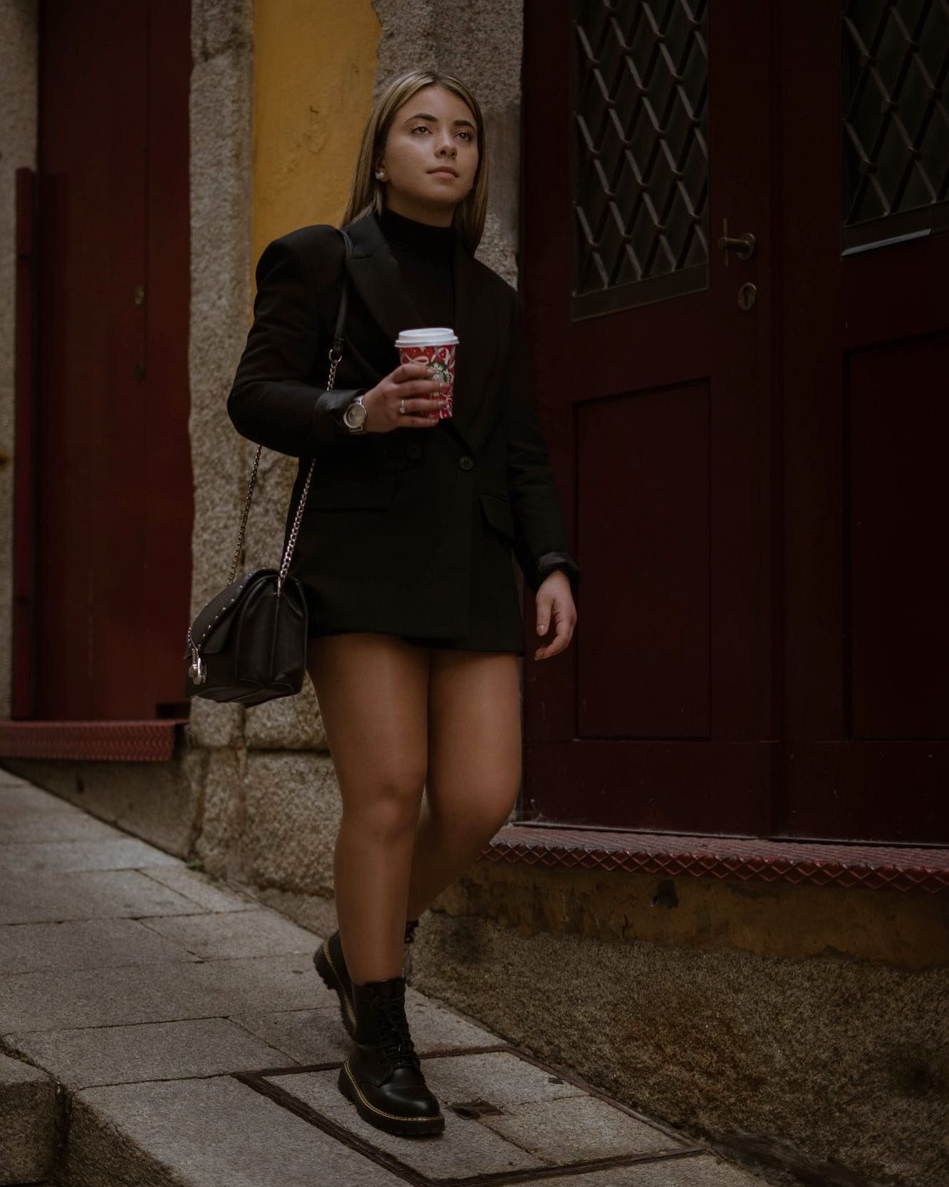 Woman in black blazer and skirt holds red coffee cup while walking on cobblestone street.