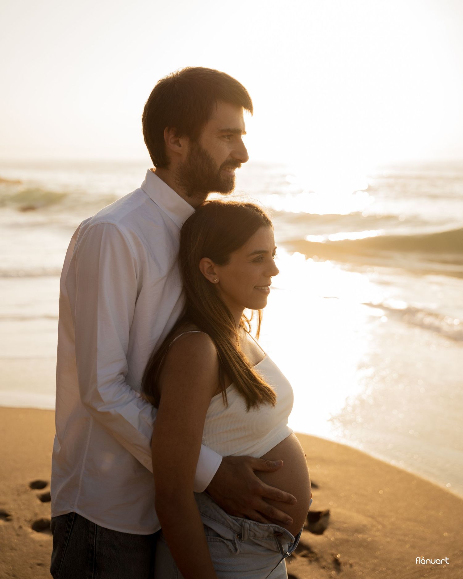 Couple embracing on a sunny beach at golden hour, man holding pregnant woman's belly near ocean waves.