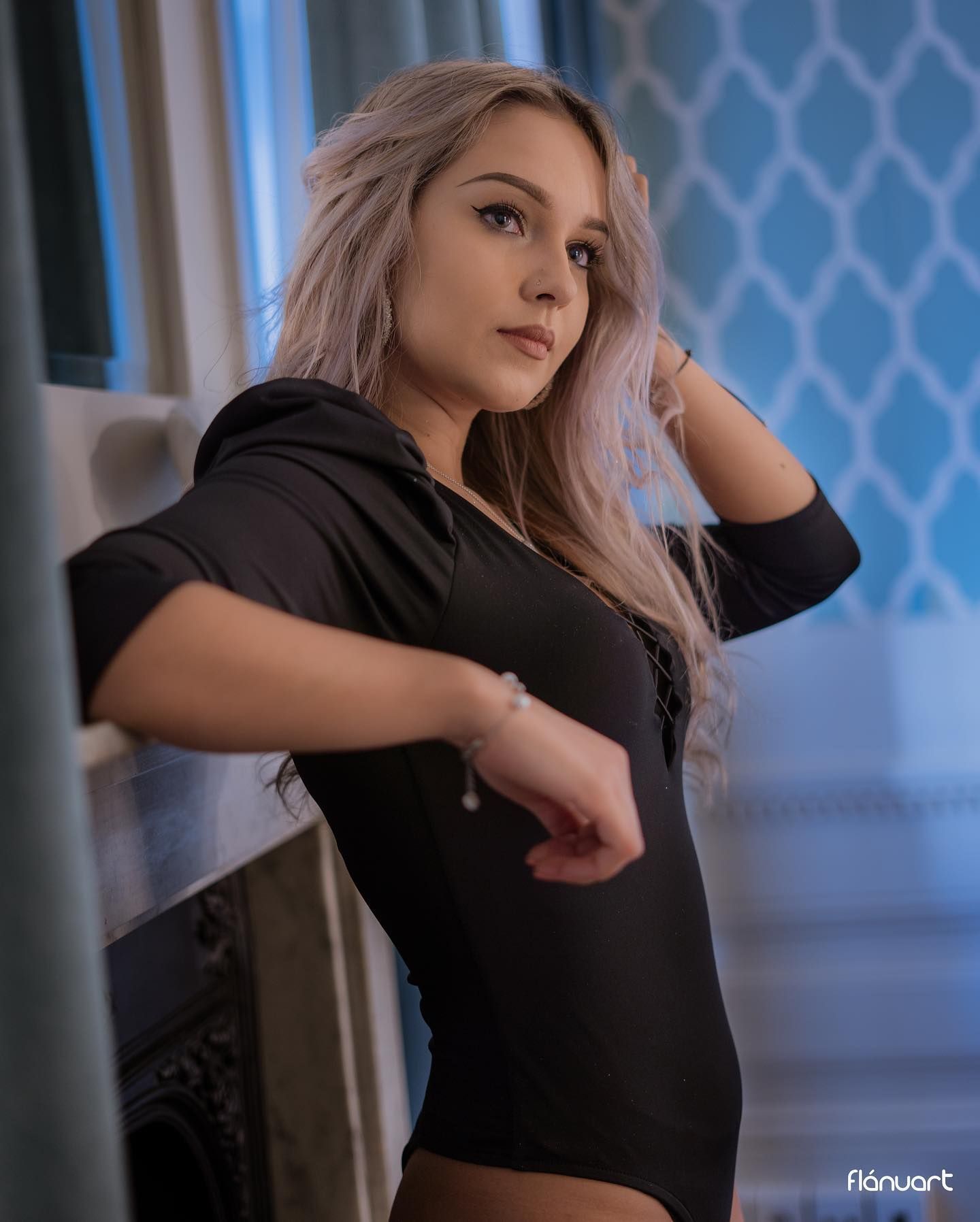 Young woman with lavender hair in black hoodie poses by window in blue room.