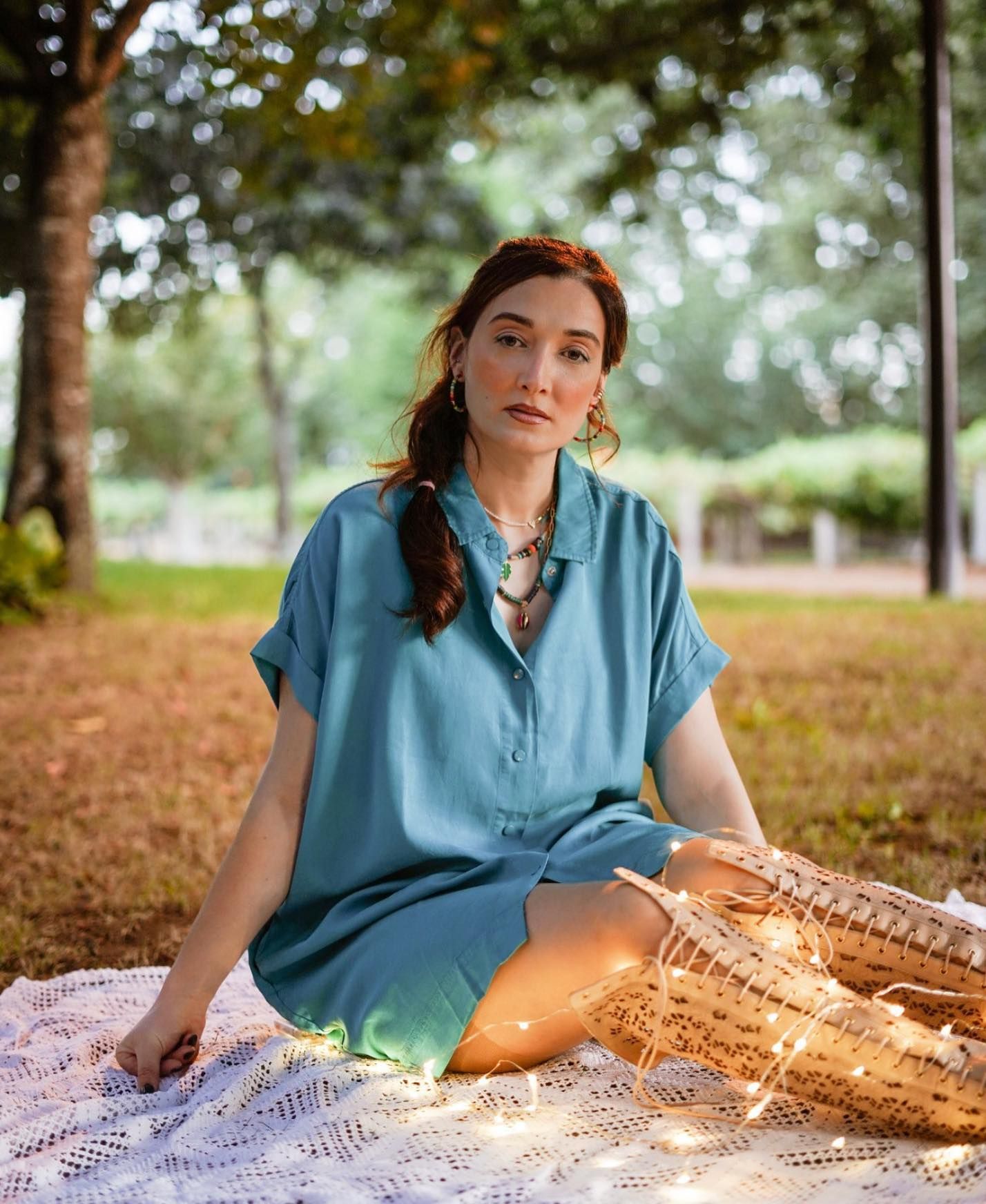 Woman in teal dress sitting on white lace blanket in park with string lights and wicker basket.
