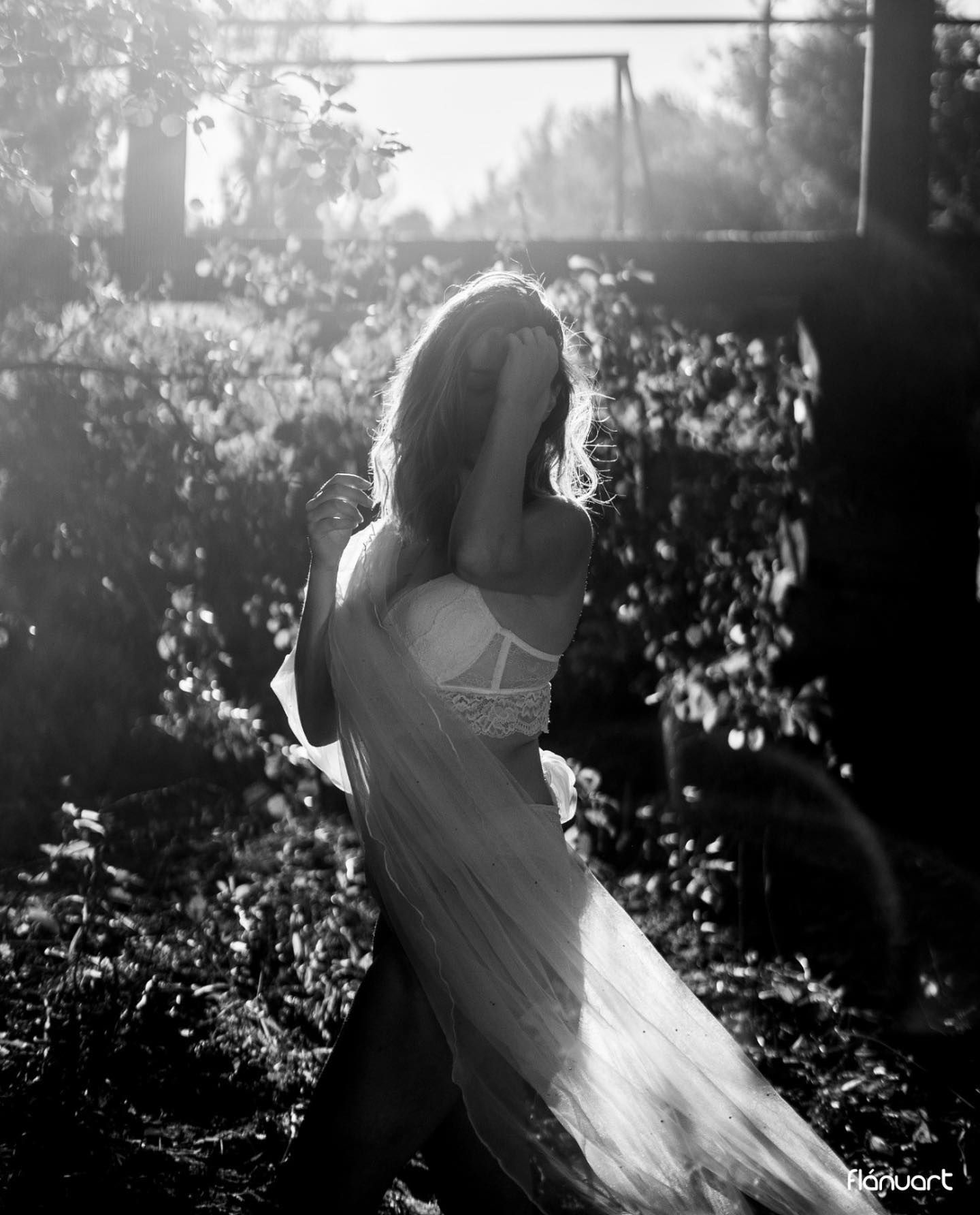 Black and white artistic photo of woman in flowing white dress in sunlit garden setting.