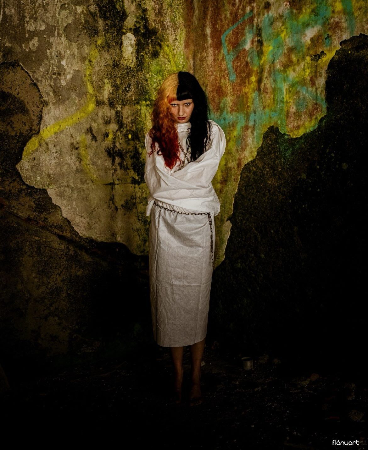 Woman in white wrapped dress standing barefoot in a dark cave with colorful textured stone walls.