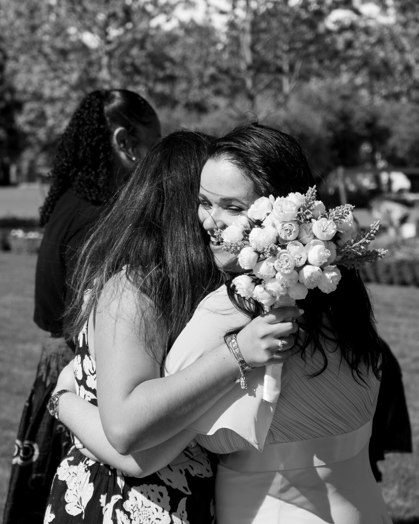 Bride in white gown joyfully embraces a guest while holding a beautiful floral bouquet outdoors.