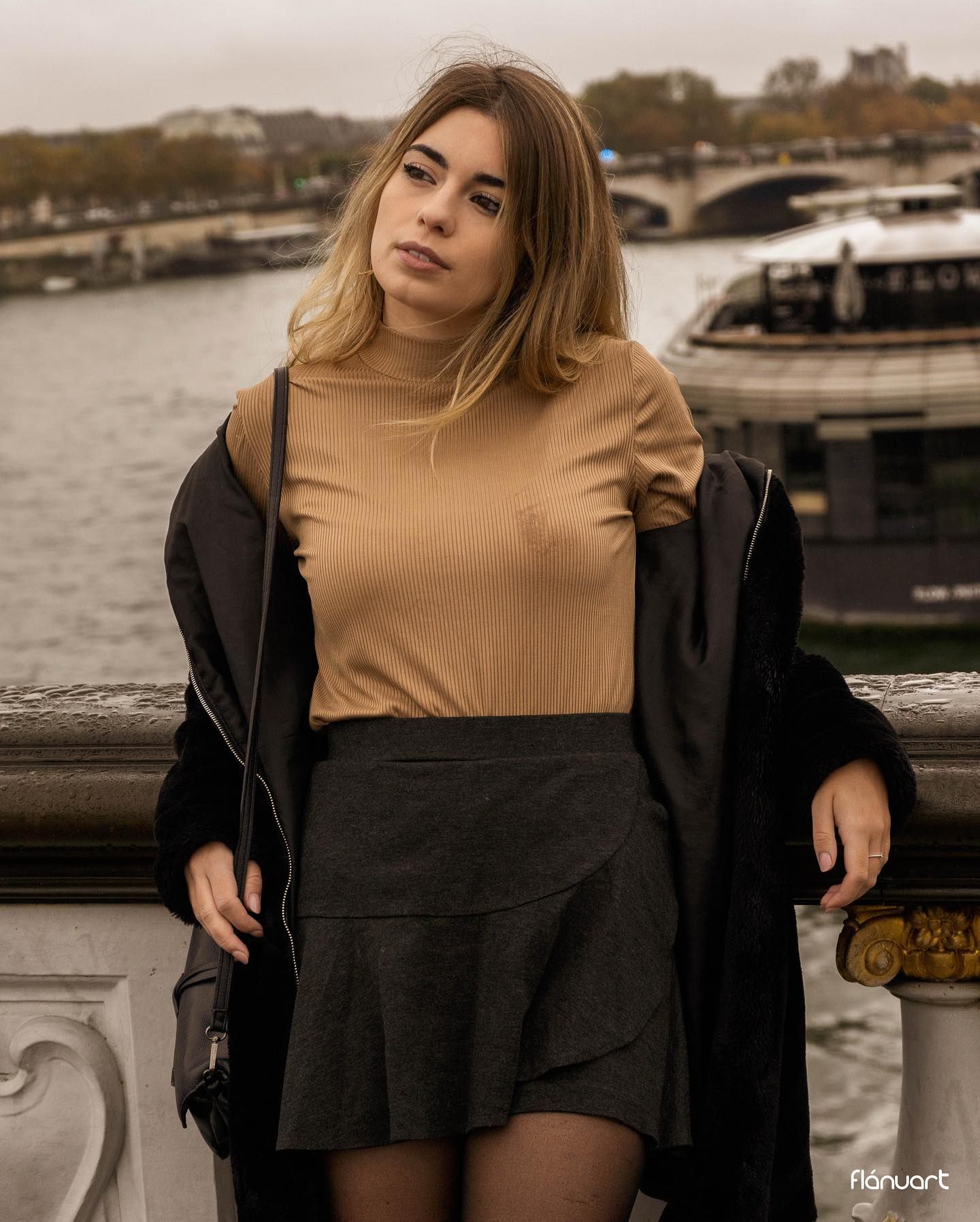 Young woman in camel top and dark skirt poses on bridge overlooking river in Paris.