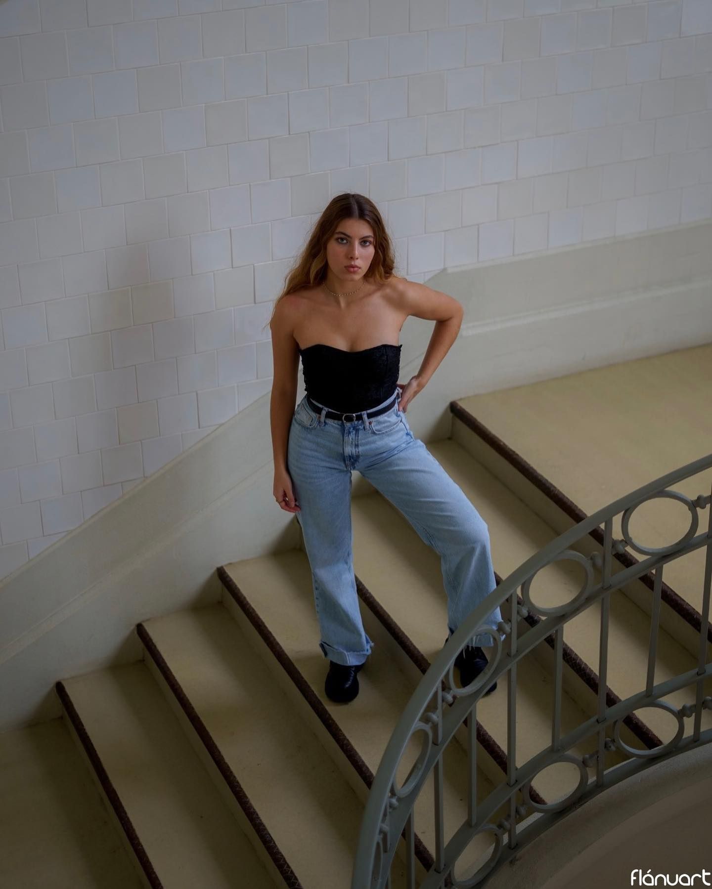 Woman in black strapless top and light jeans posing on a staircase with curved railing.