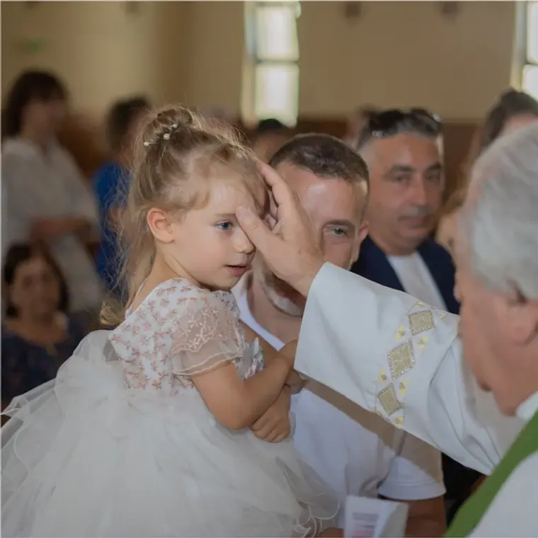 A priest blesses a young girl in a white dress during a Catholic baptism ceremony in a church.