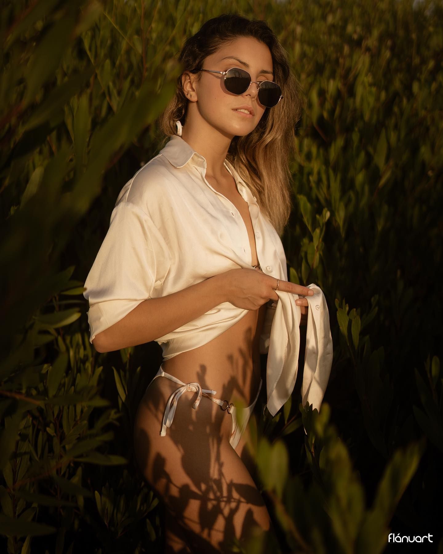Woman in white tied shirt and sunglasses posing among tall green plants at sunset.