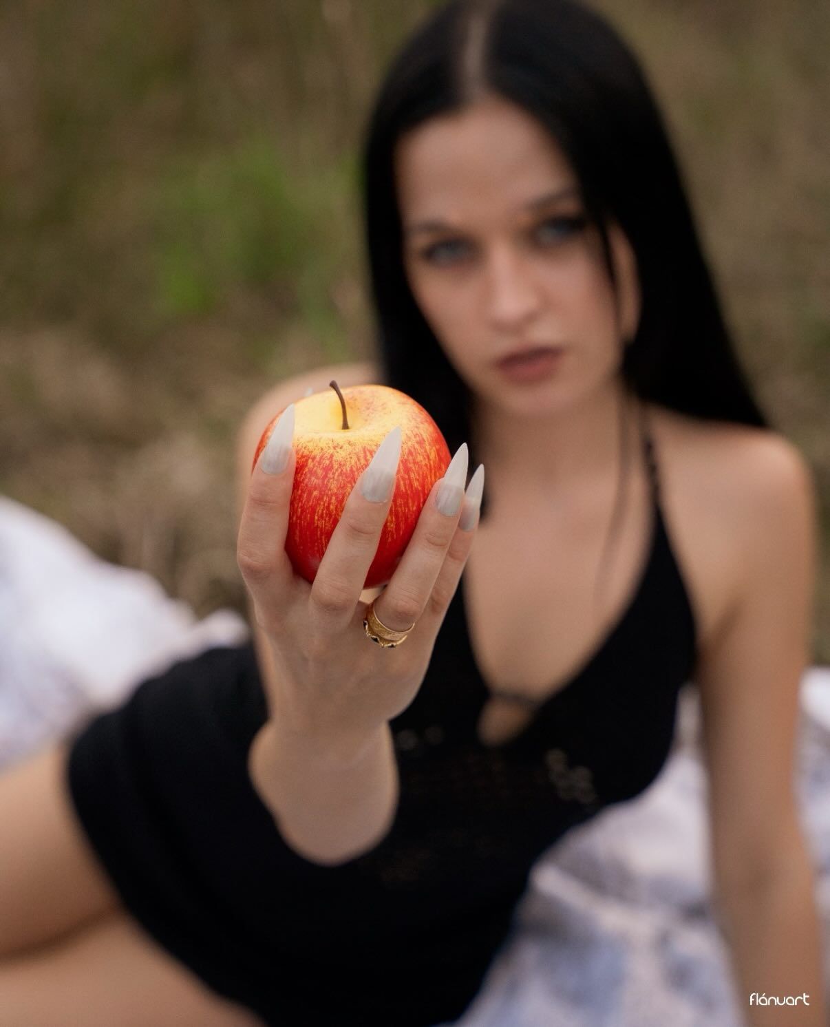 Dark-haired woman holding red apple toward camera while reclining outdoors in natural setting.