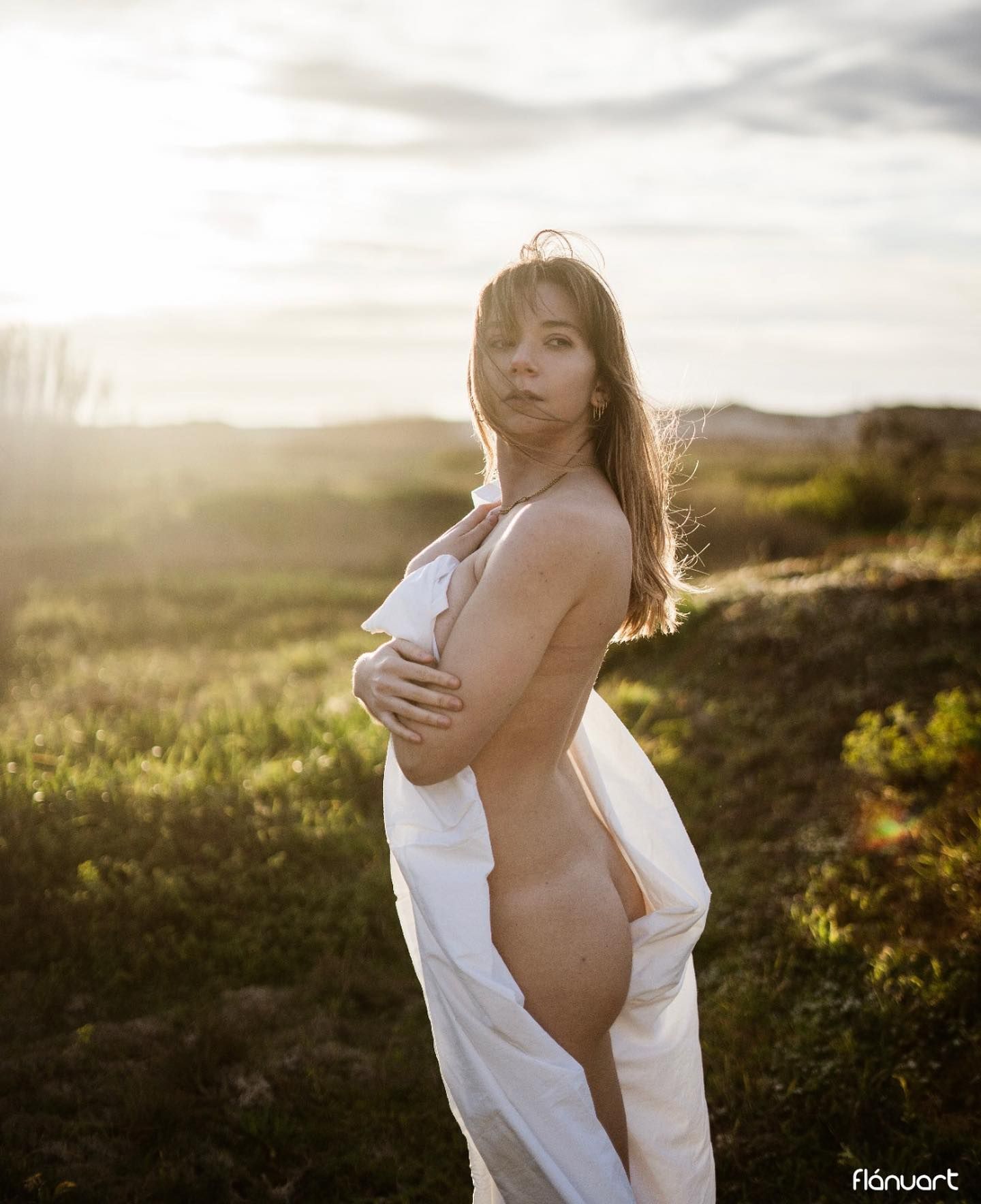 Woman draped in white fabric posing outdoors in golden sunset light over open landscape.