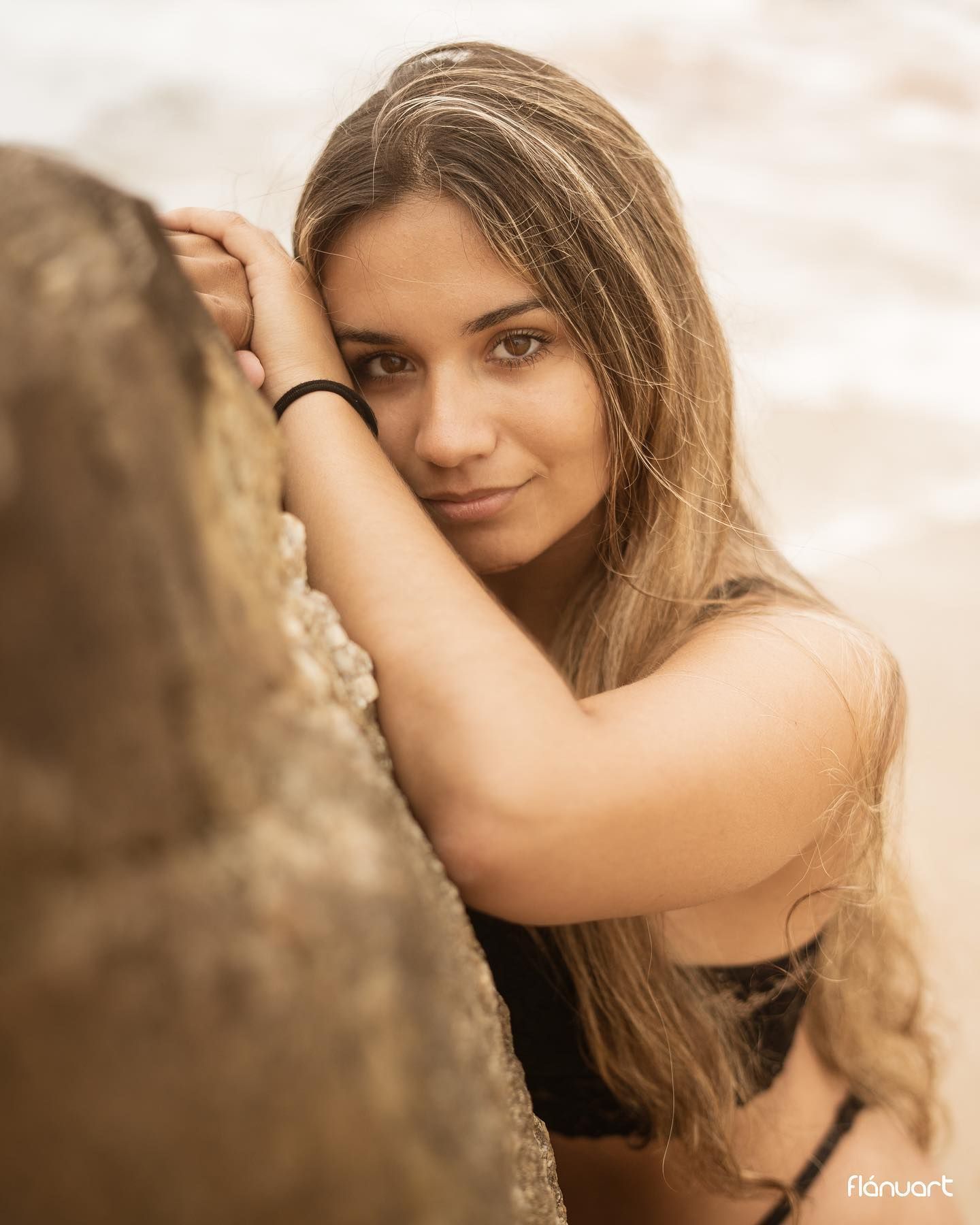 Young woman with long blonde hair leaning against a rock near the ocean.