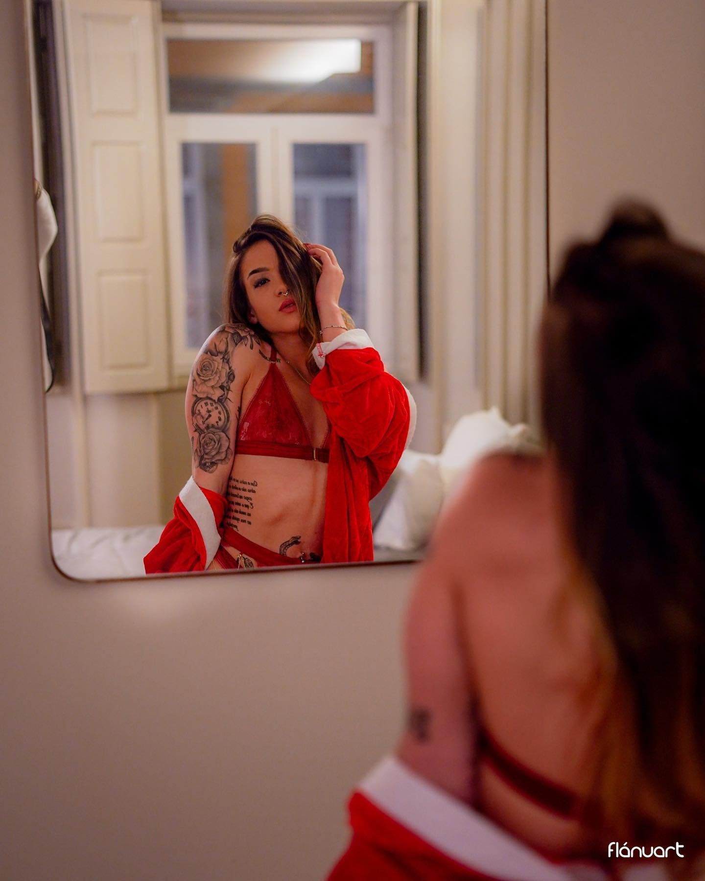 Tattooed woman in red lingerie viewing her reflection in a bedroom mirror.
