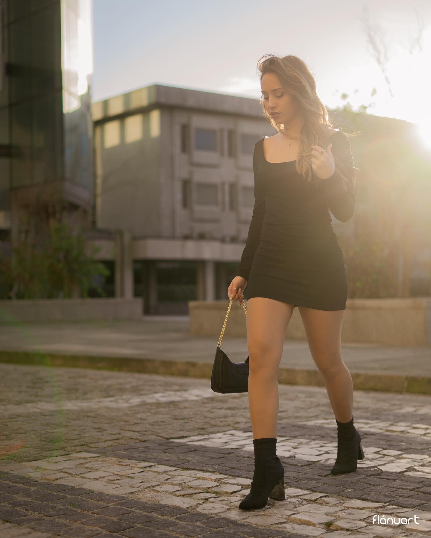 Woman in black mini dress and ankle boots walks on cobblestone plaza at sunset.