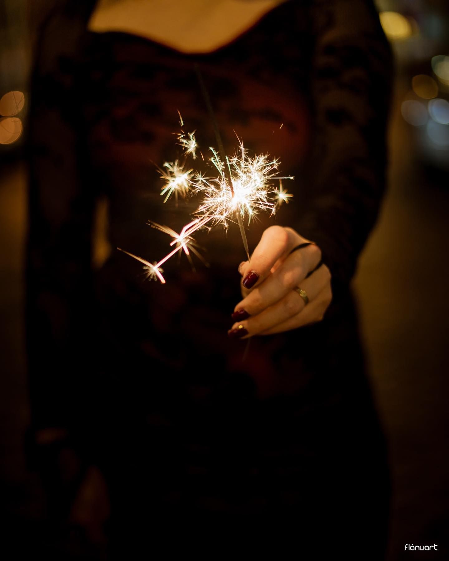Woman in black lace dress holding glowing sparkler in dark nighttime setting.
