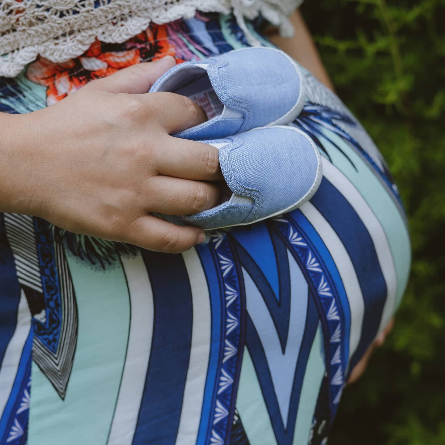 Close-up of pregnant woman in colorful dress holding tiny blue baby shoes against her belly.
