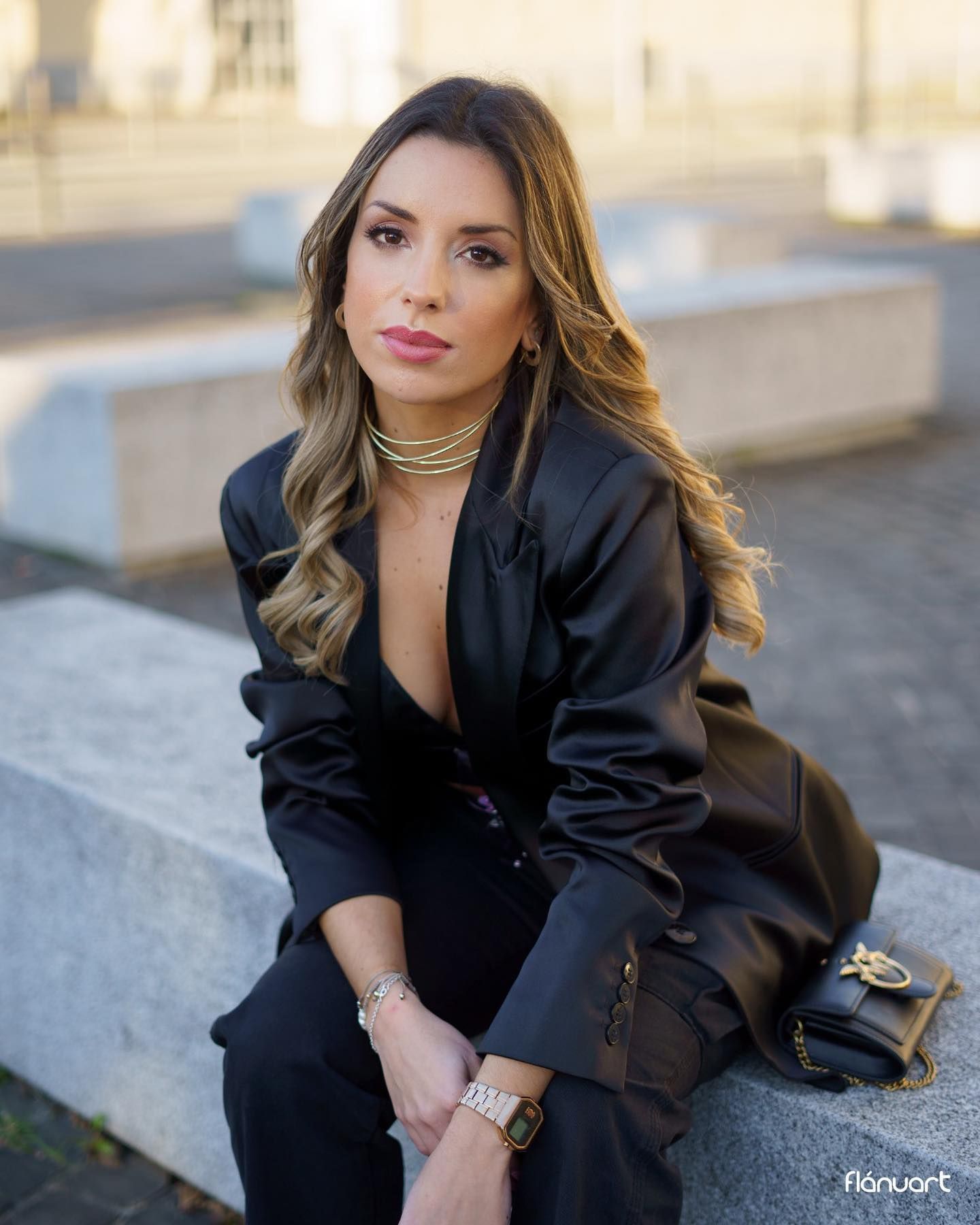 Elegant woman in black satin blazer with gold jewelry and designer bag sitting on a stone bench outdoors.